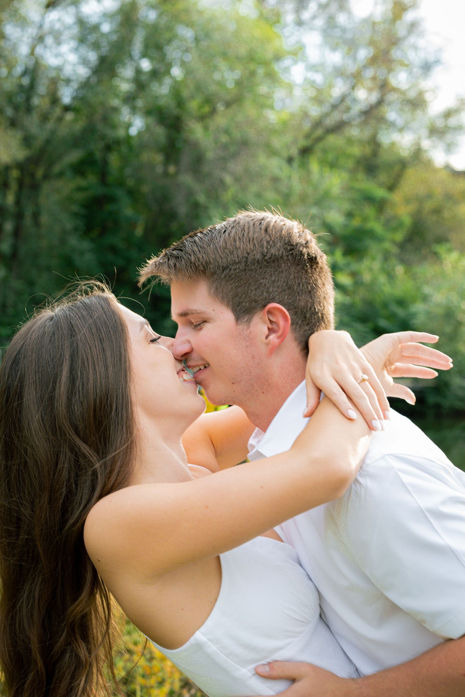 Couple sharing a romantic kiss outdoors, woman wearing engagement ring, embracing in front of lush green trees.