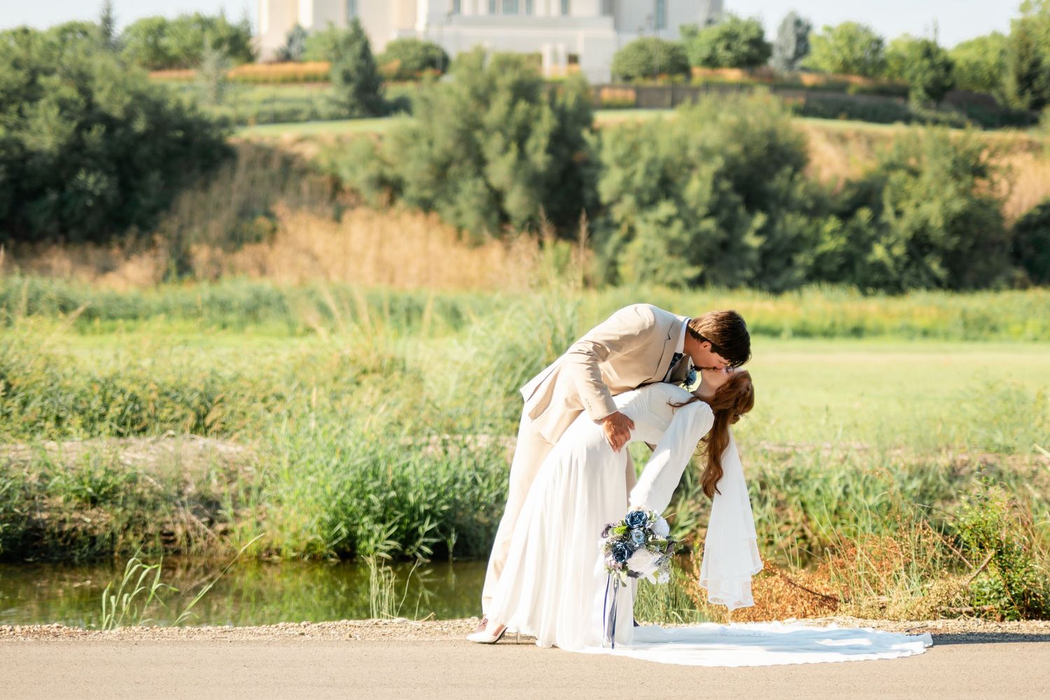 Bride and groom share a romantic dip kiss near a scenic pond with lush greenery and a white temple in the background.