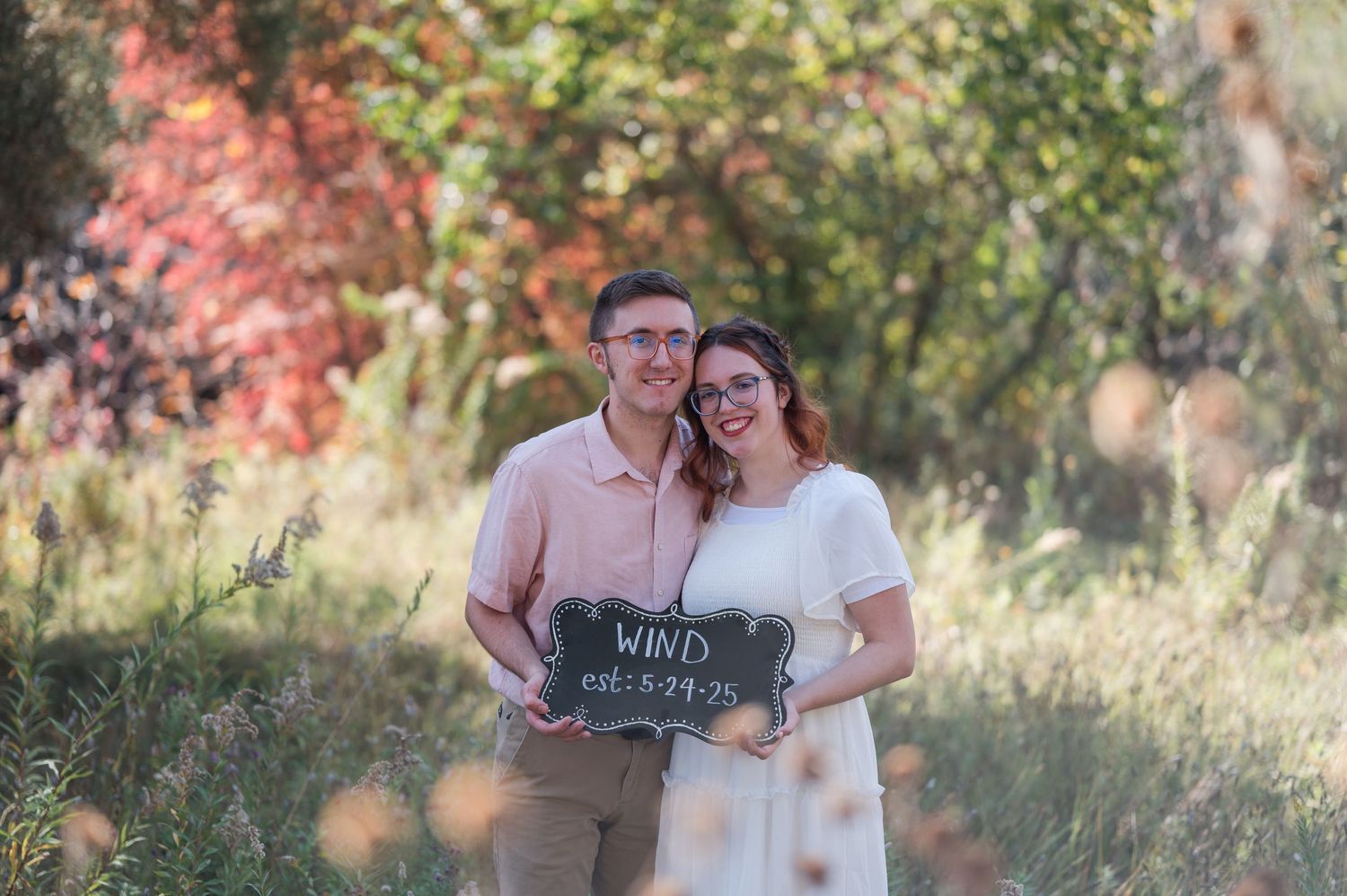 Couple holding a chalkboard wedding sign in a colorful autumn outdoor setting.