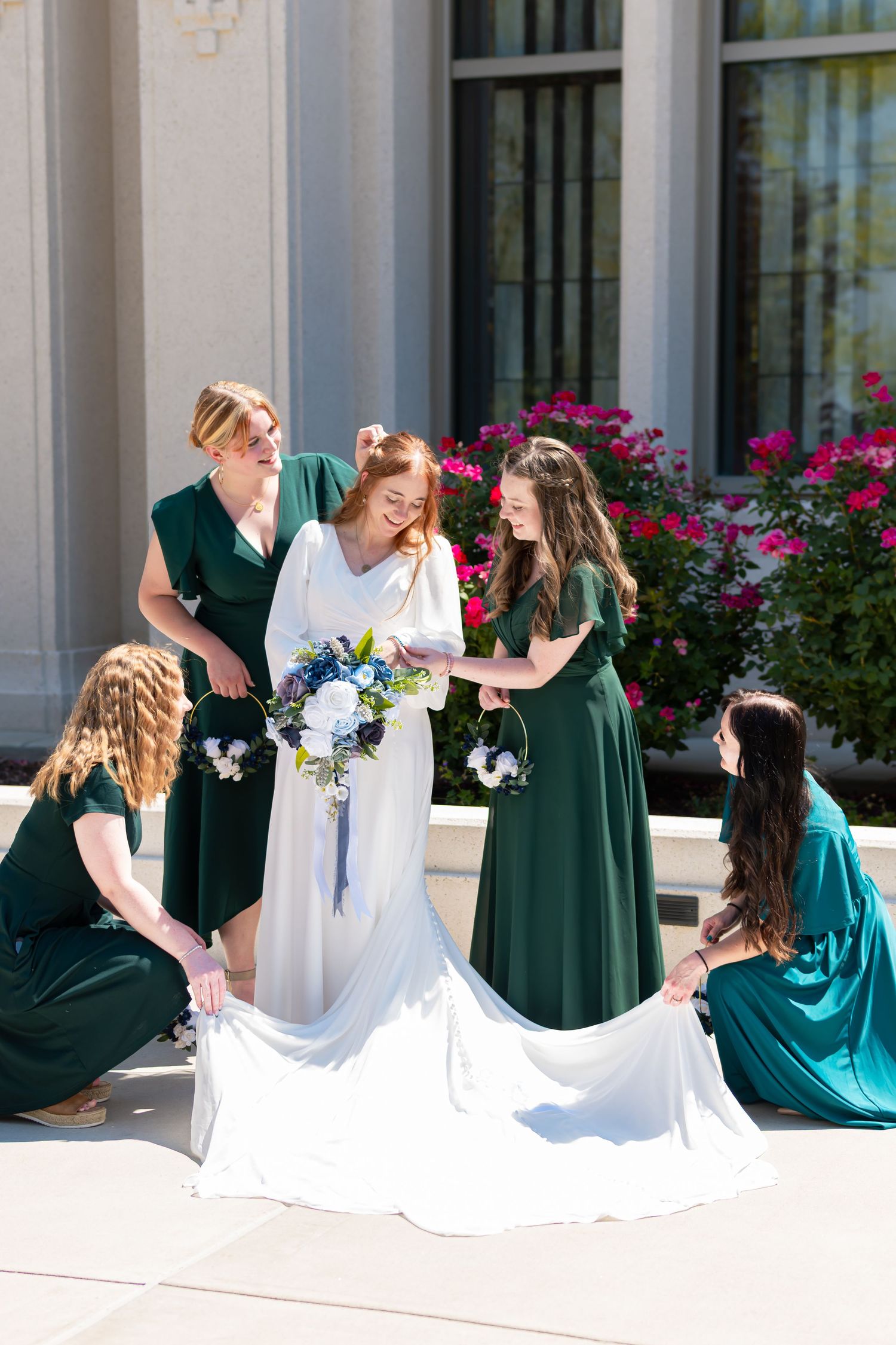 Bride in white gown holds bouquet surrounded by bridesmaids in green dresses adjusting her train outdoors.