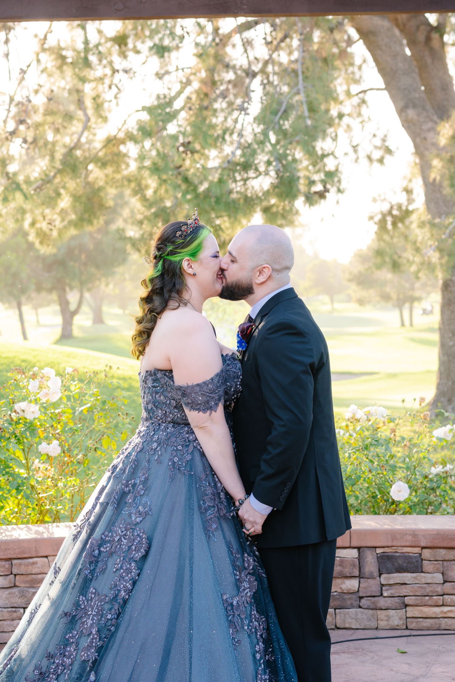 Bride in gray floral gown and groom in black suit share a kiss outdoors at golden hour wedding ceremony.