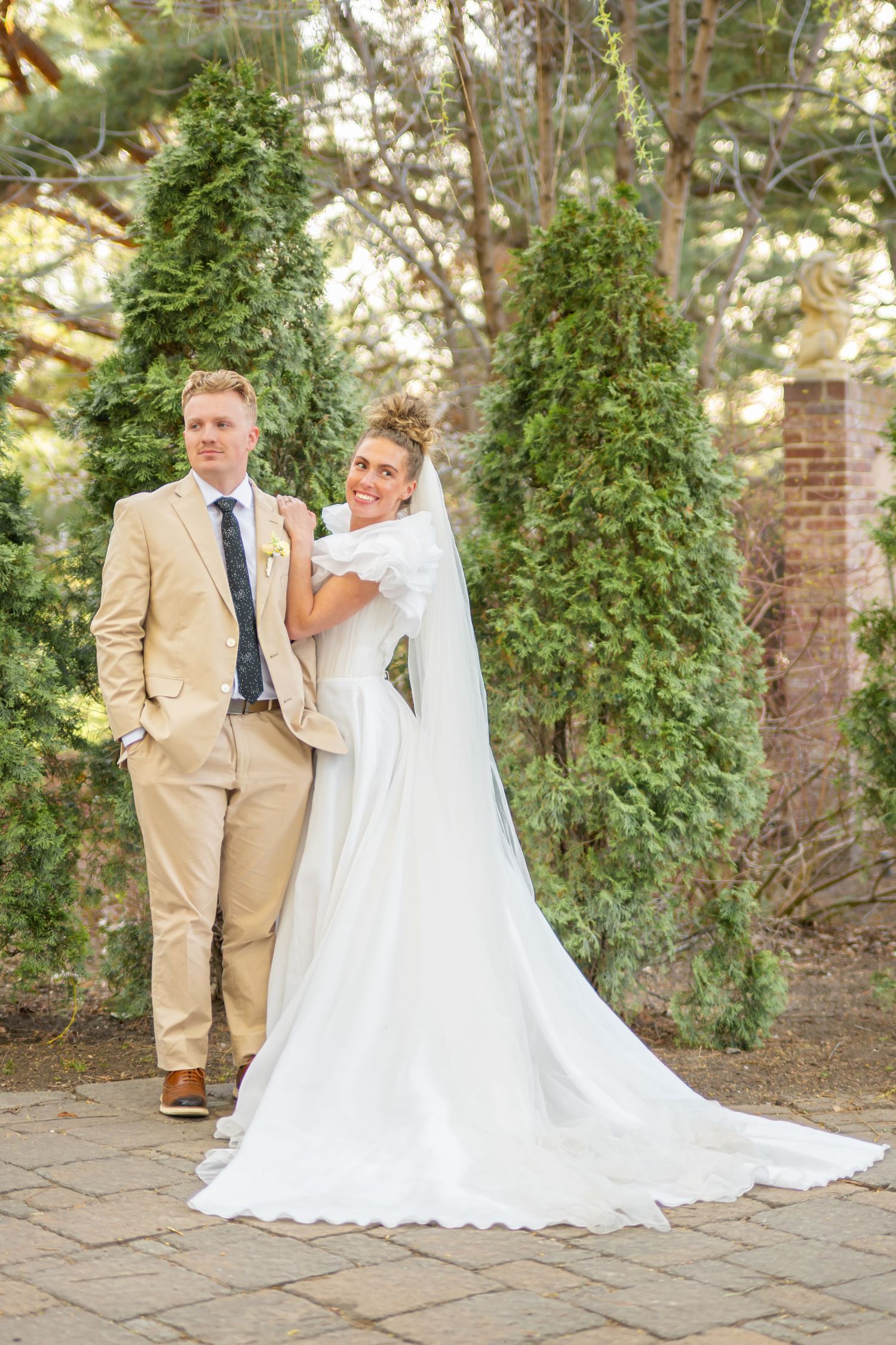 Bride in a white gown with a long veil and groom in a tan suit pose together on a stone patio surrounded by evergreen trees.
