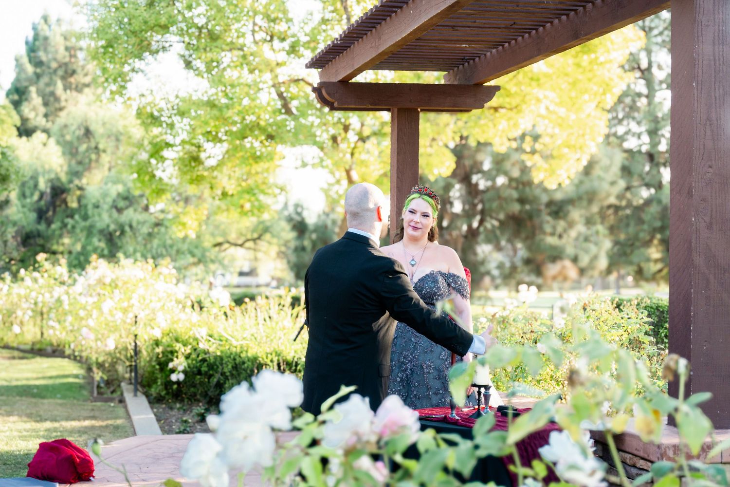 Couple sharing a romantic moment under a wooden pergola surrounded by lush greenery and white flowers in a garden setting.