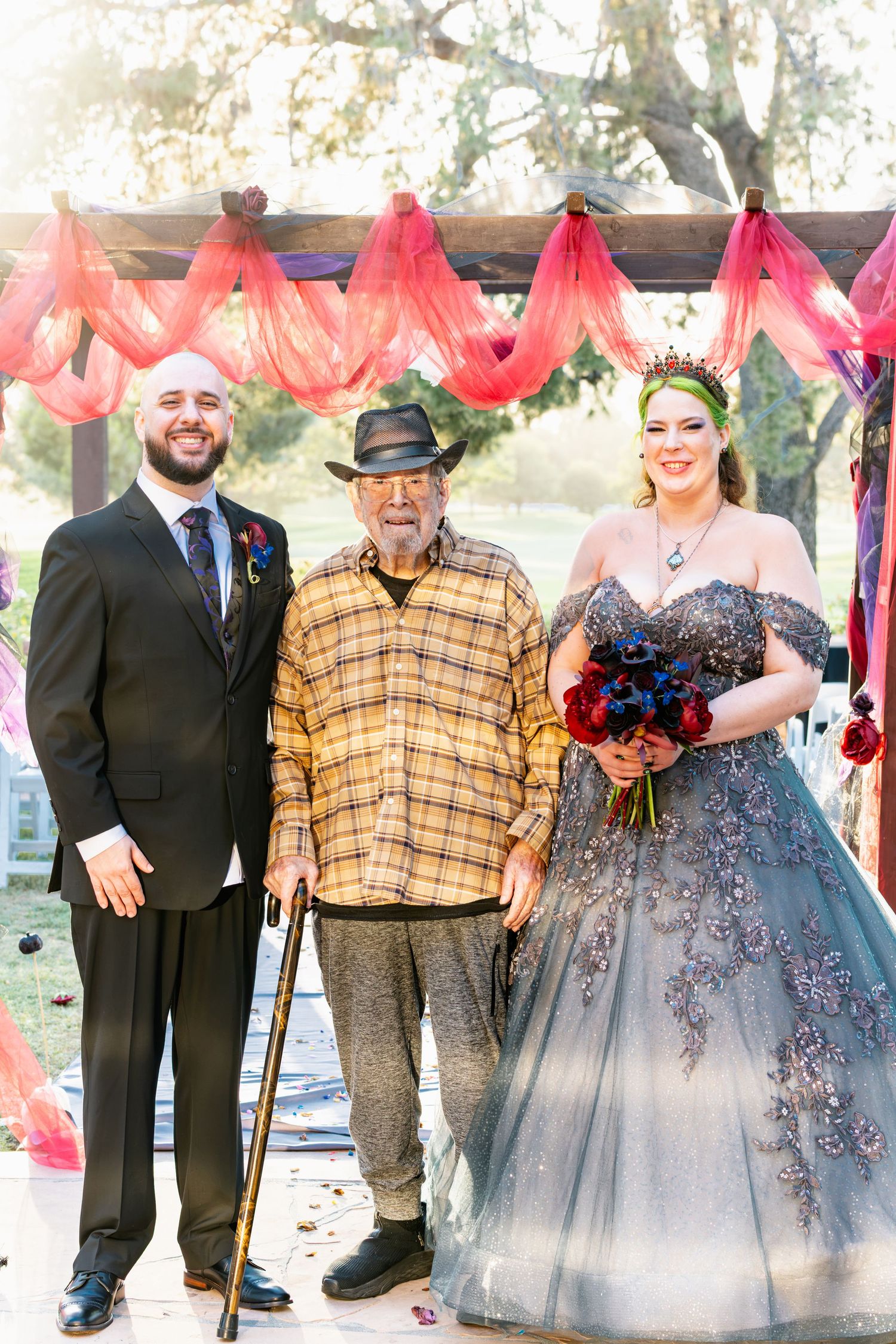 Bride in blue gown and groom in black suit pose with elderly man using cane at outdoor wedding ceremony.