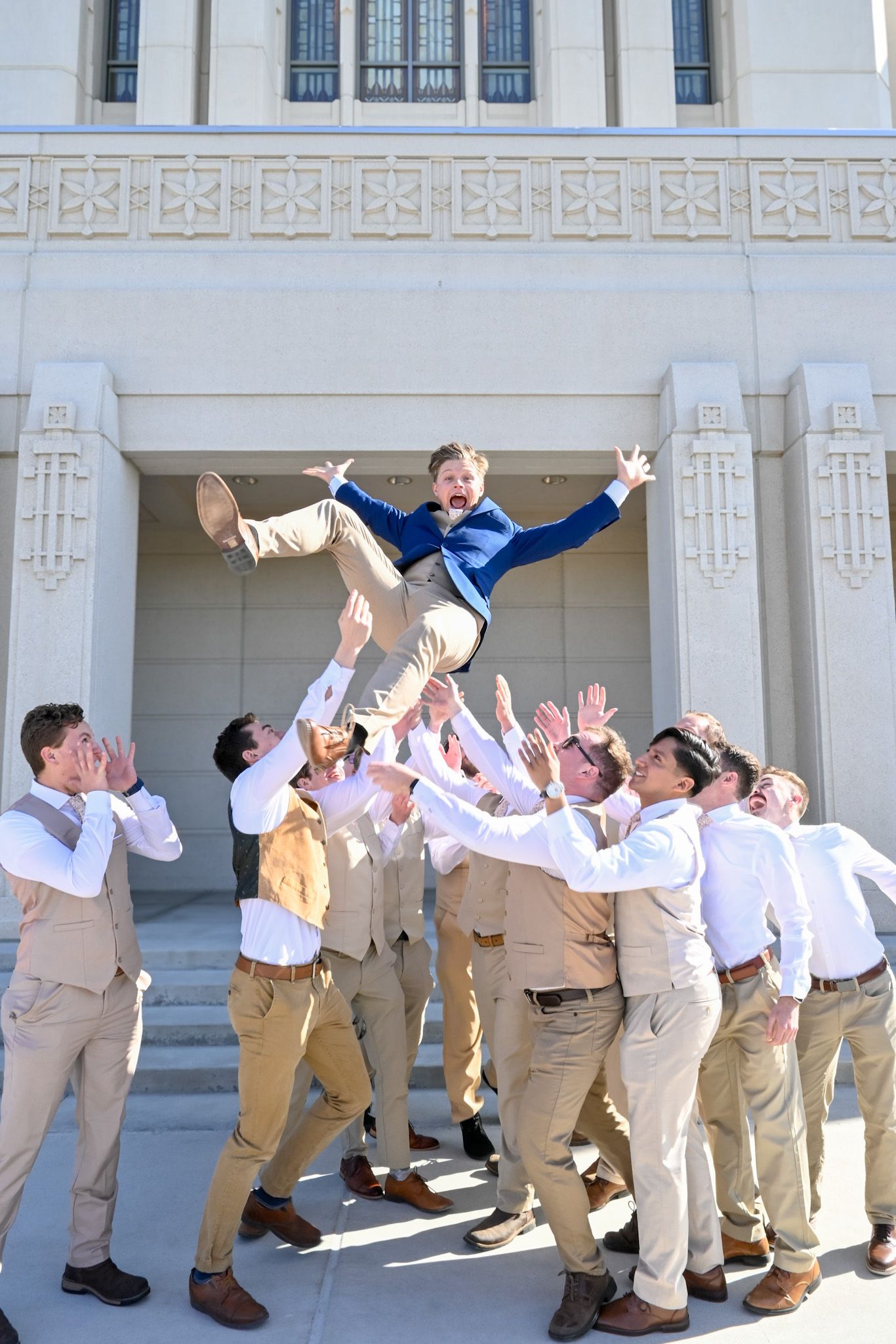 Groomsmen in khaki pants and white shirts playfully tossing groom in the air outside church building.