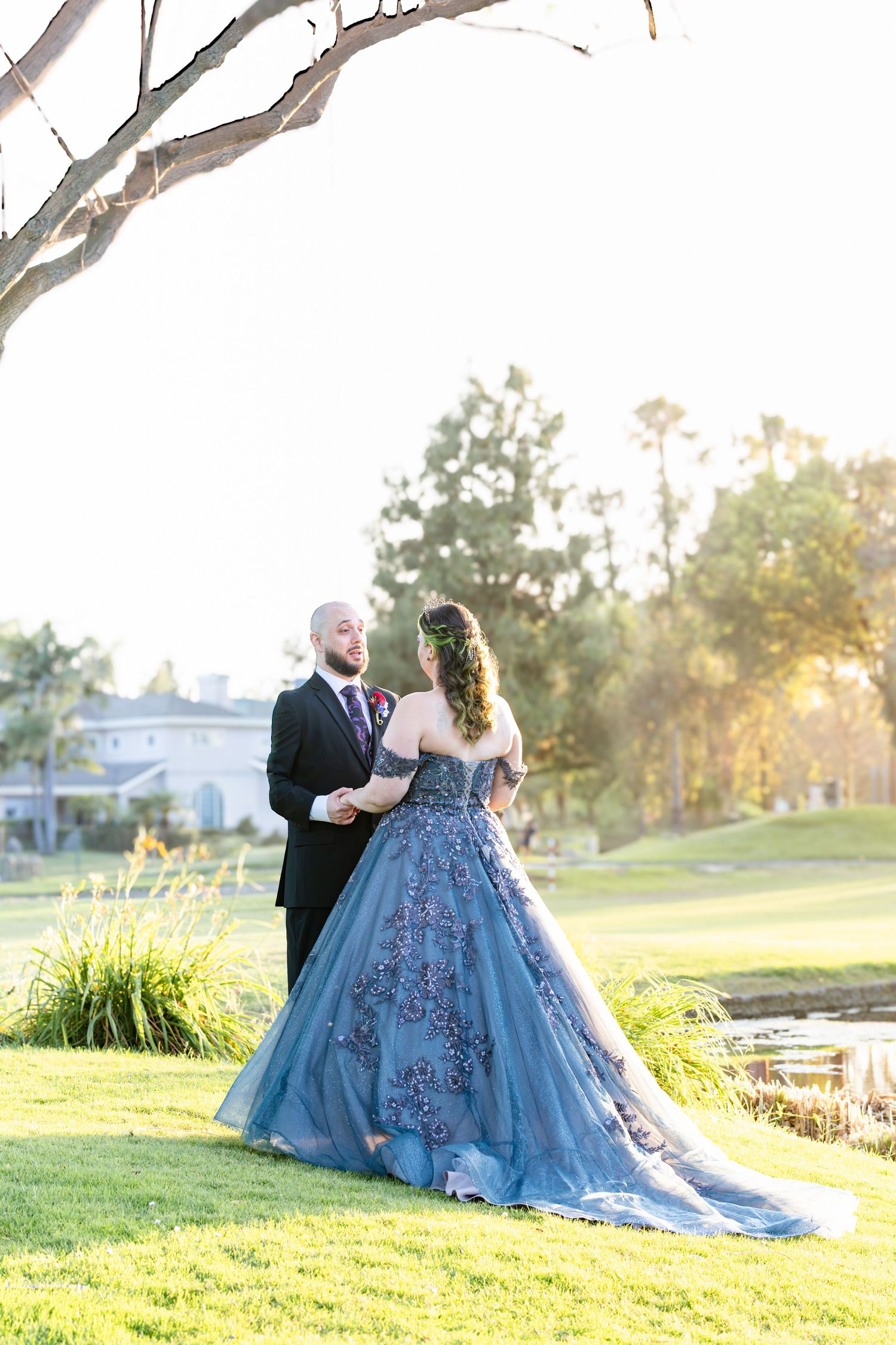 Couple in elegant wedding attire, blue floral gown and black tuxedo, share a romantic moment on a lush green lawn.