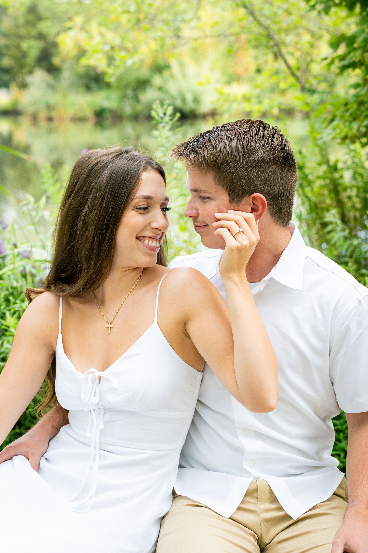 A happy couple in white outfits sit together outdoors near a pond, sharing a romantic moment surrounded by lush greenery.