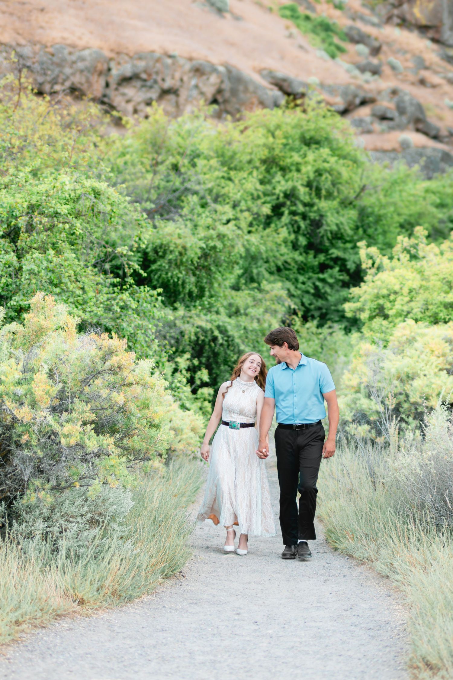 Couple holding hands and smiling at each other while walking down a gravel path lined with green shrubs and rocky hills.