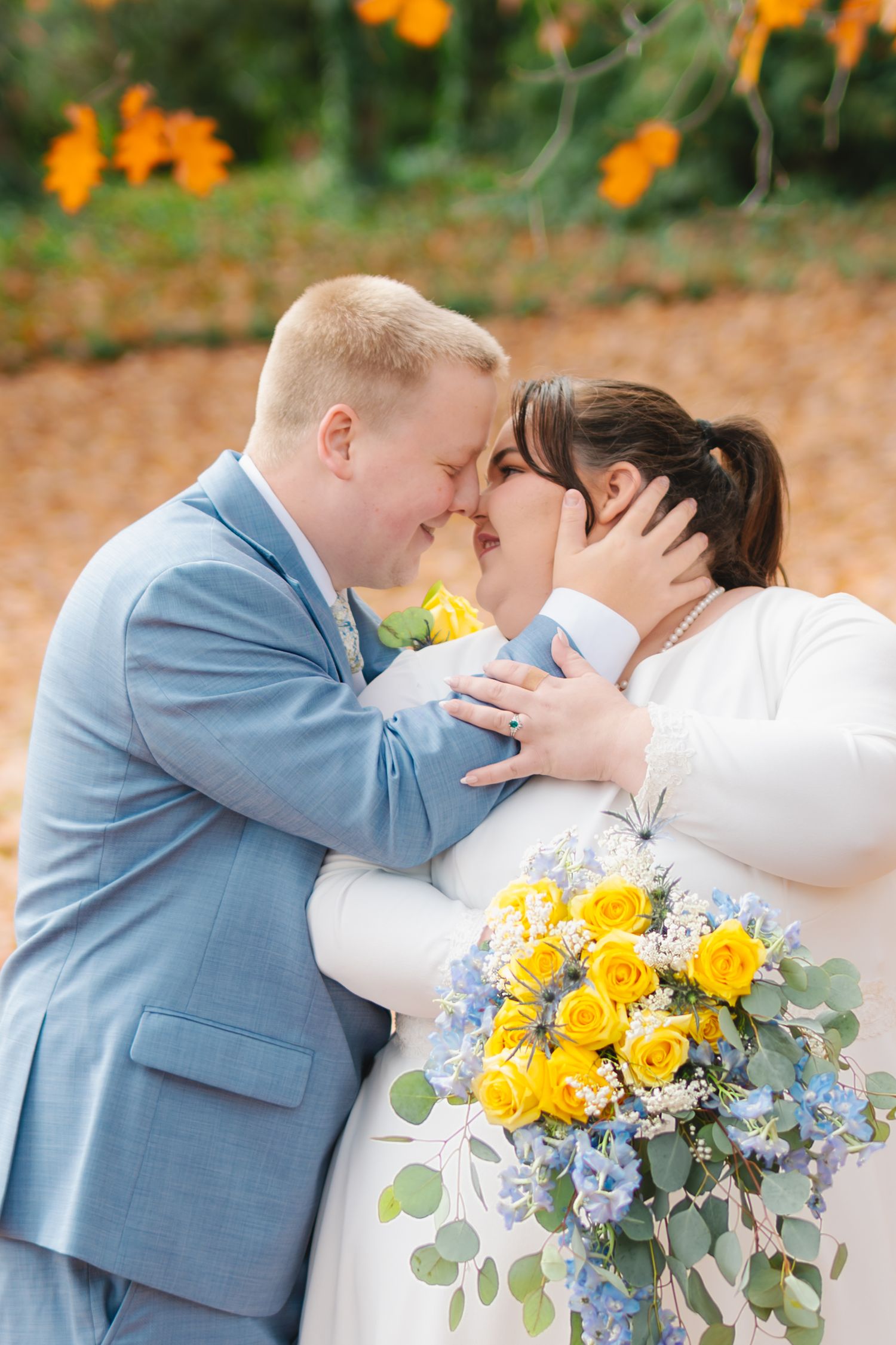 A romantic wedding moment captured outdoors with a blue suit and white dress, featuring a bright yellow rose bouquet.