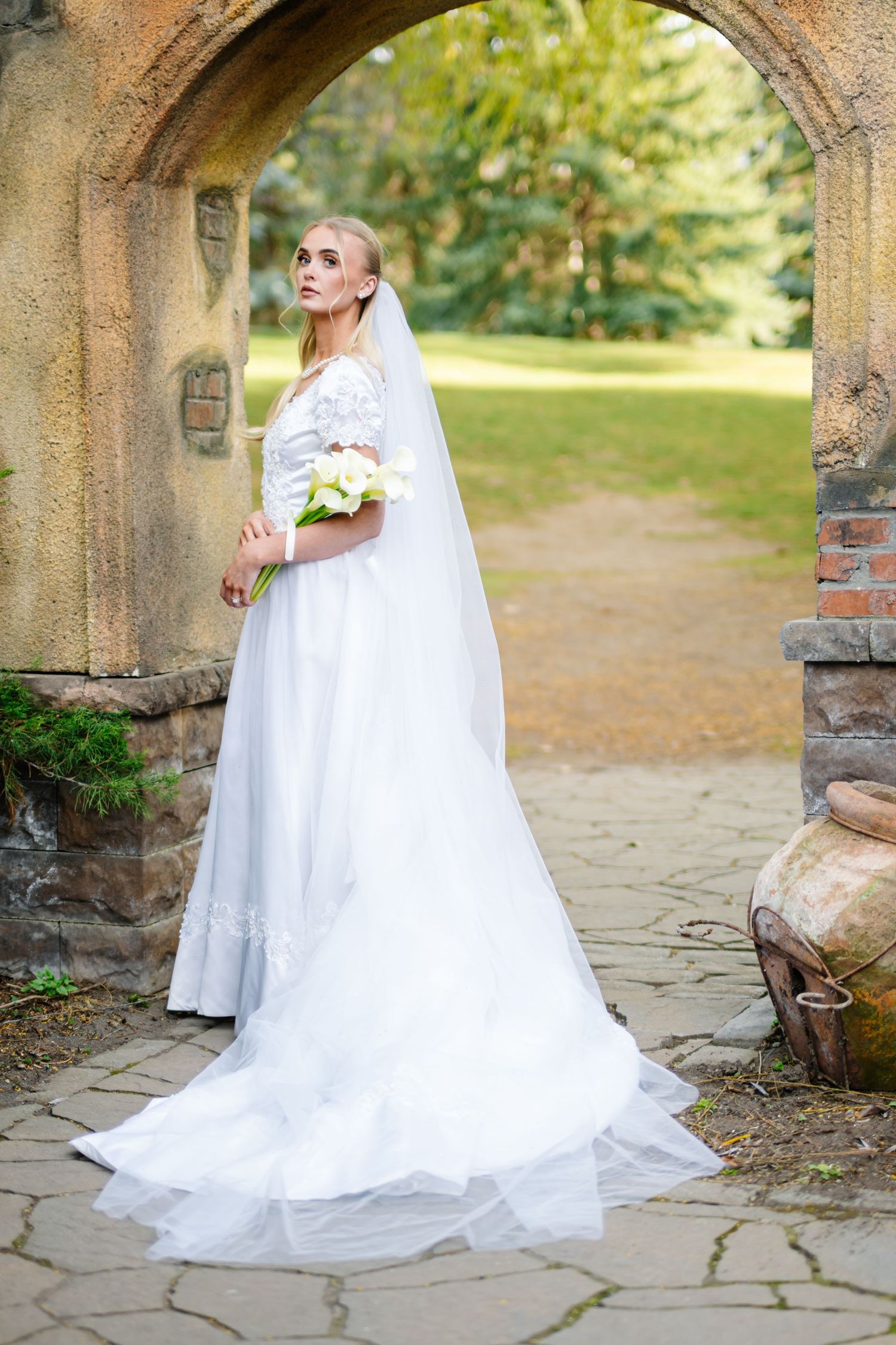 A woman in a long white wedding dress stands in an archway surrounded by greenery and brick walls.