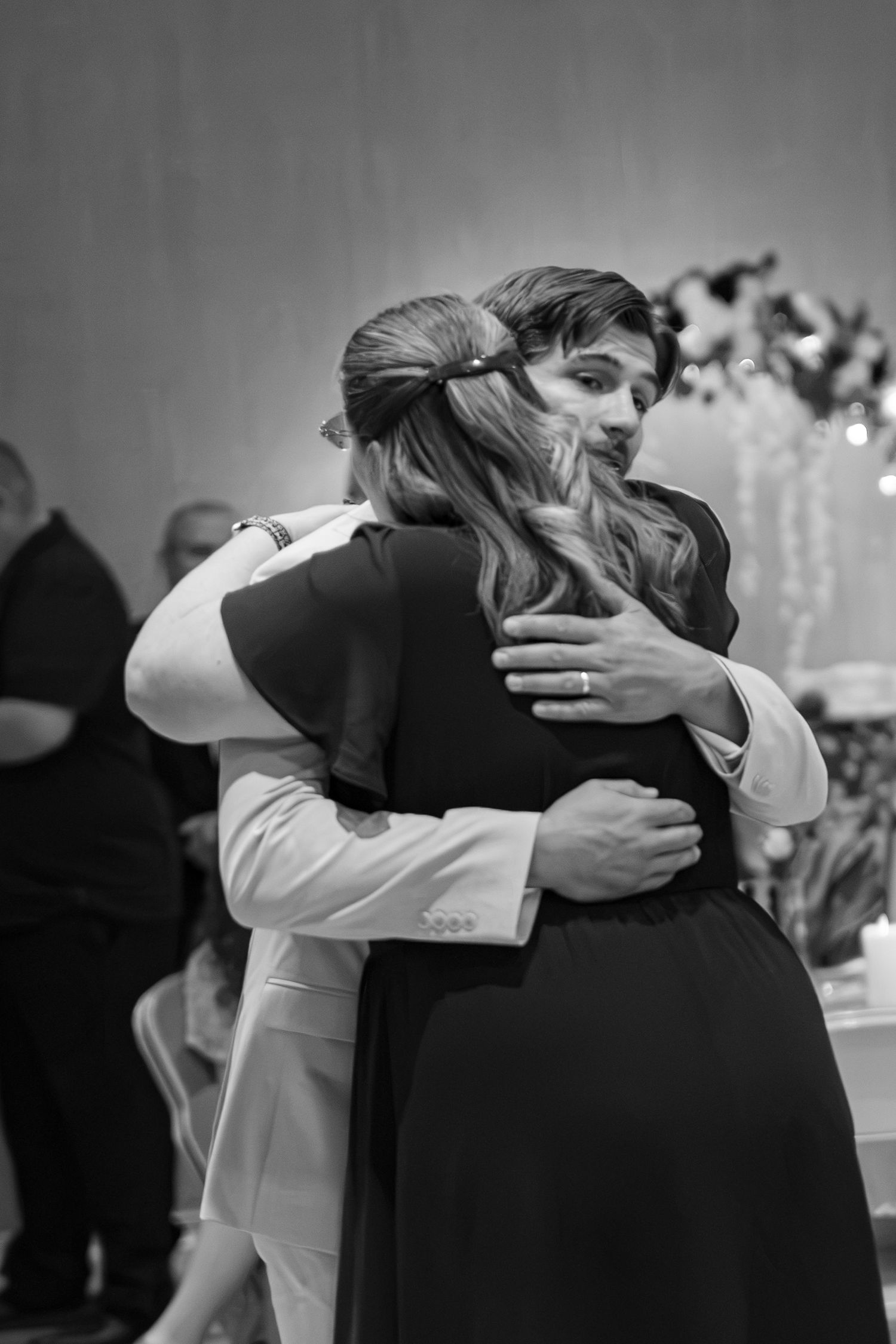 Black and white photo of groom sharing a heartfelt hug with a guest at wedding reception.