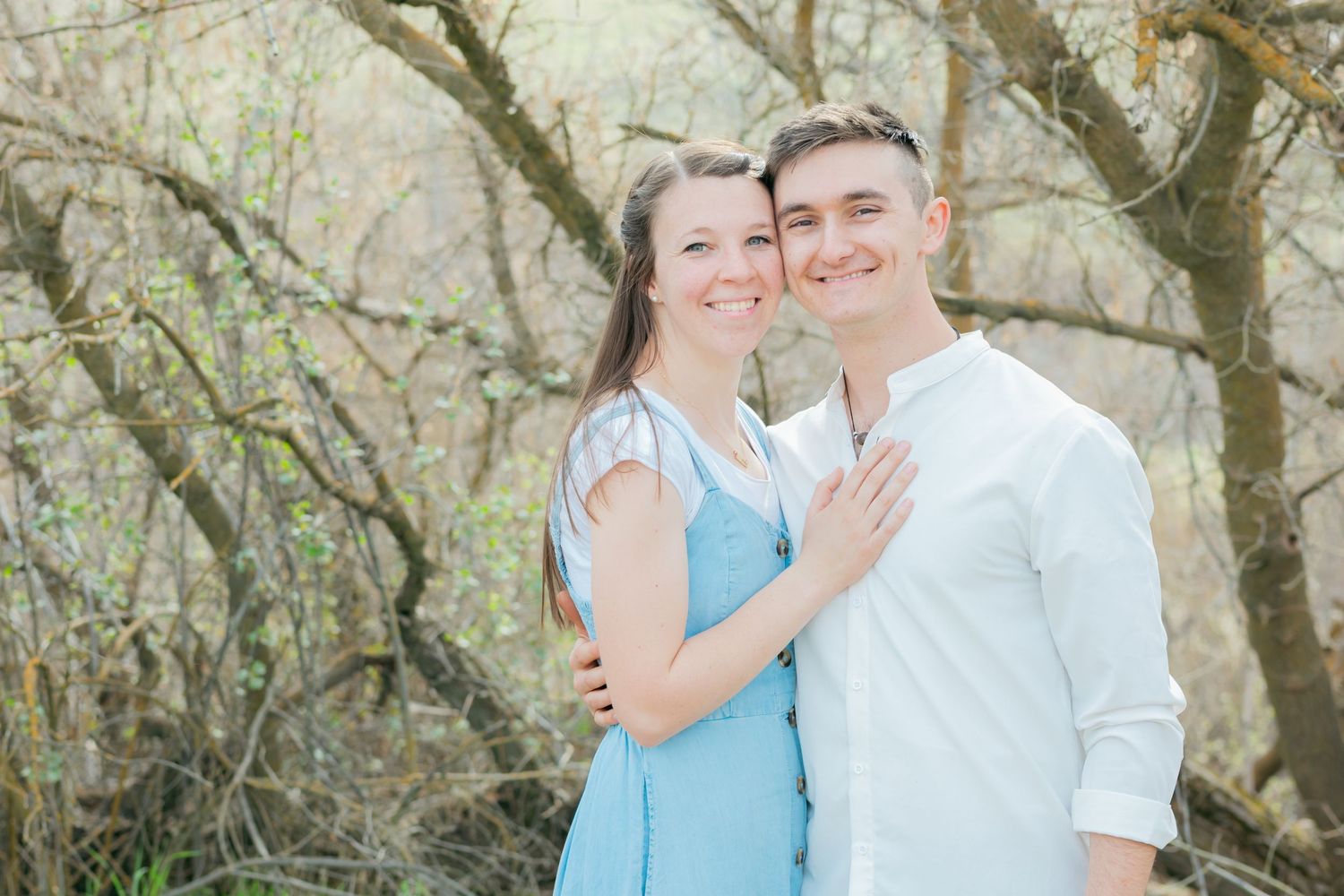 A smiling couple embraces outdoors among bare trees, woman in blue dress, man in white shirt, soft natural light.