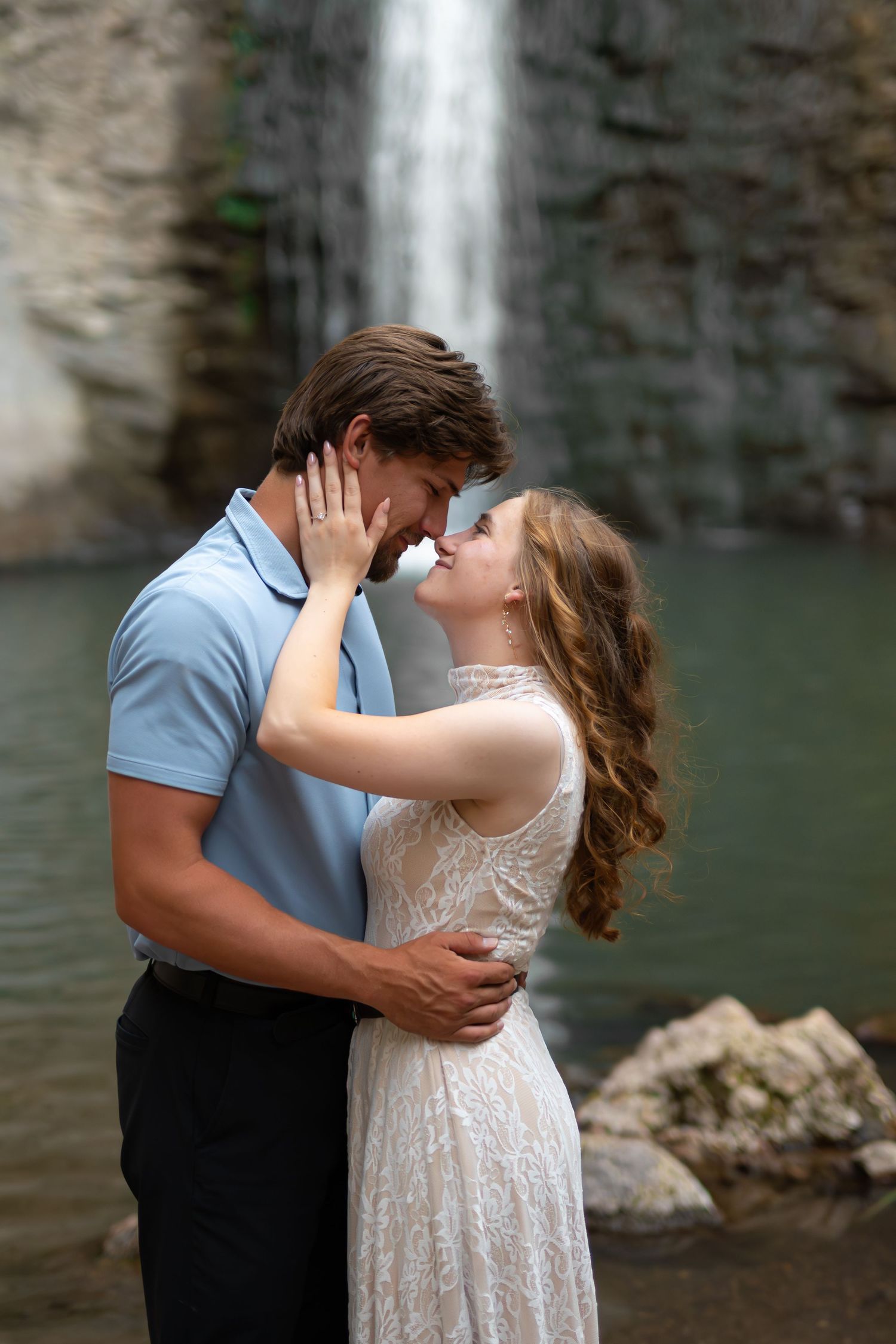 A couple shares a romantic moment in front of a waterfall, the woman in lace dress and man in blue polo embracing.