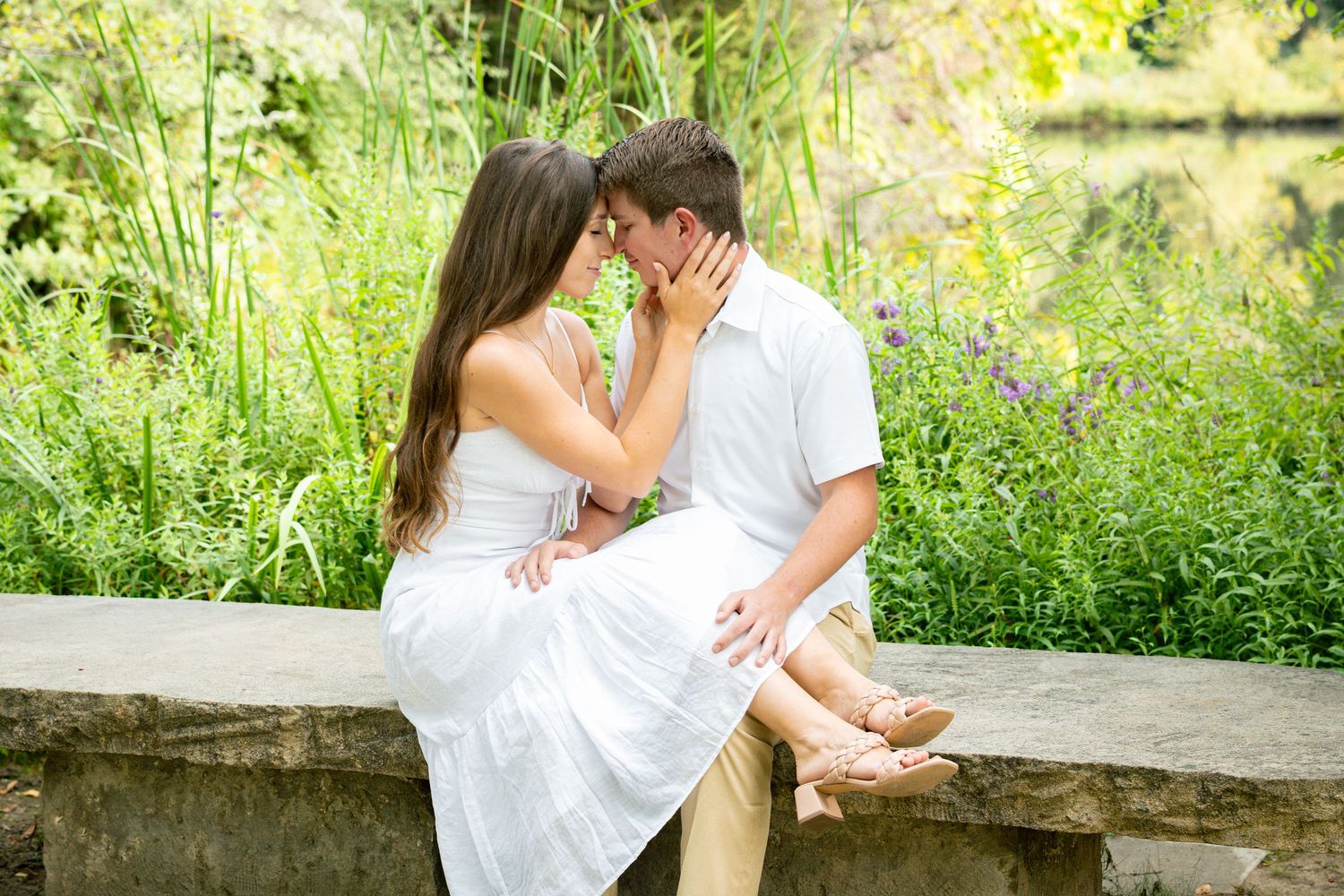 A couple shares a romantic moment sitting on a stone wall surrounded by lush green gardens and wildflowers.