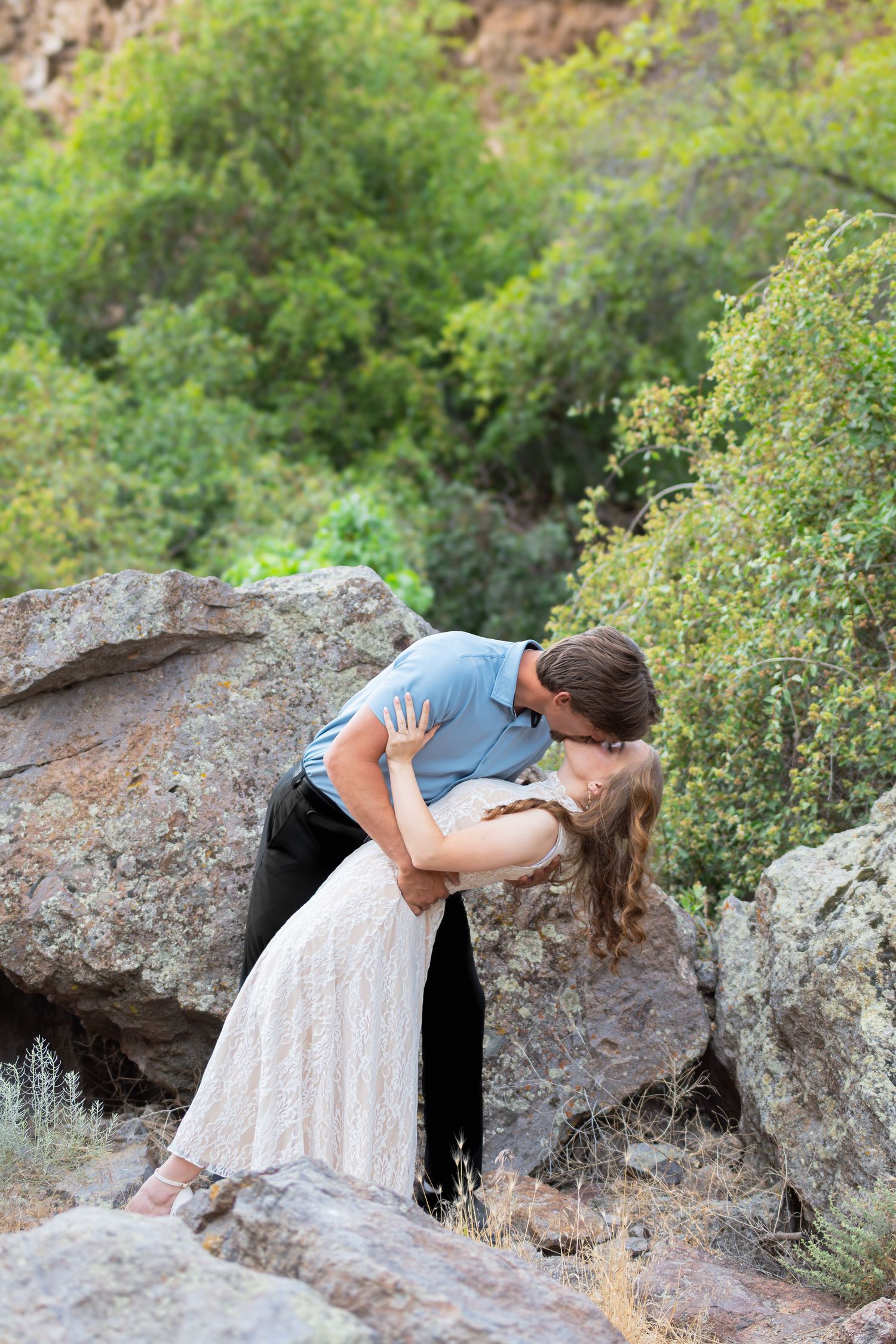Couple sharing a romantic dip kiss among large rocks with lush green trees in background.