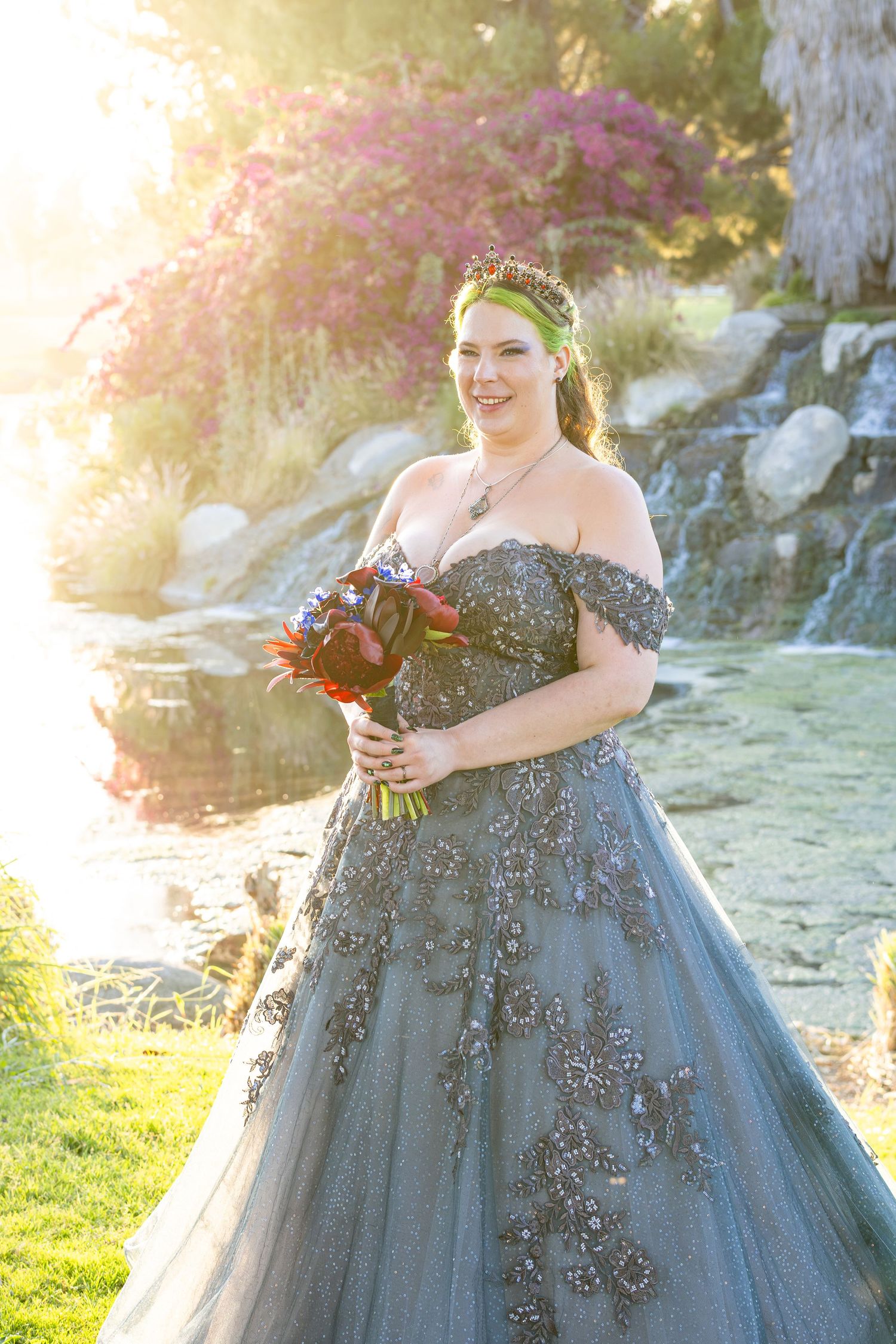 Bride in gray beaded off-shoulder ball gown with floral crown holds bouquet by waterfall in garden.