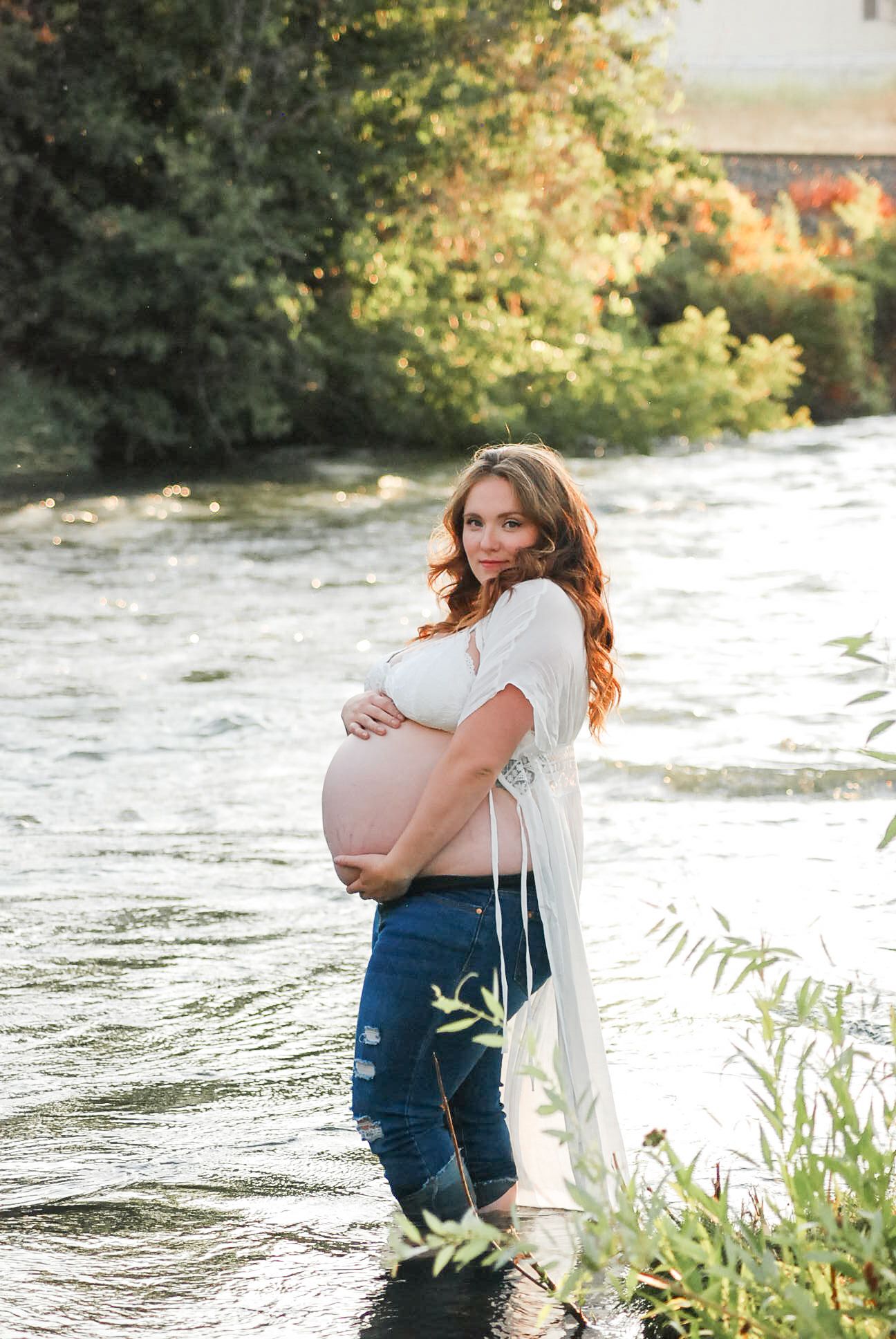 An expectant mother in white stands near flowing water and greenery during a sunset photoshoot.