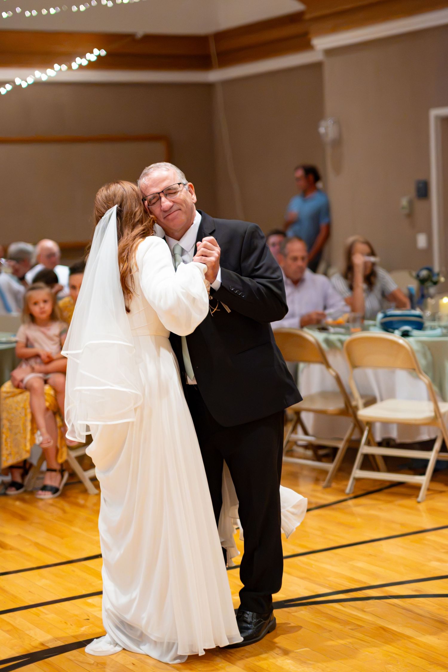 Bride and father share an emotional first dance at wedding reception on hardwood floor with guests watching in background.