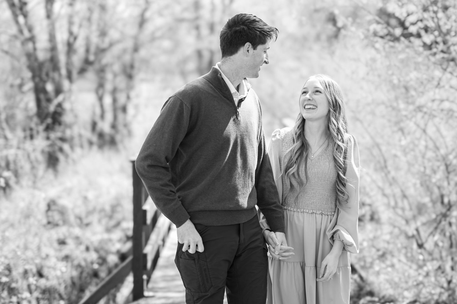 Black and white photo of a smiling couple holding hands and walking together on a bridge surrounded by trees.