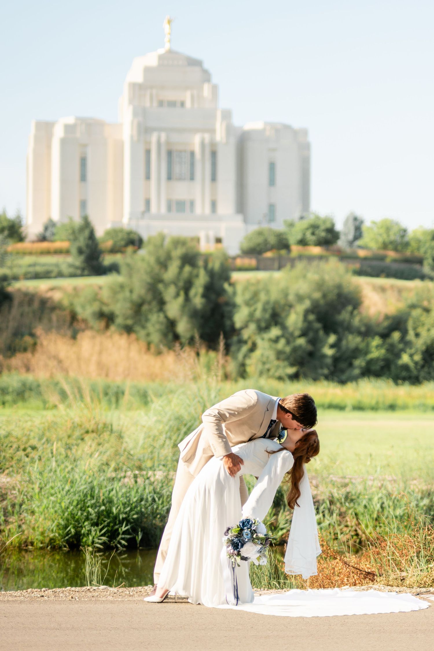 Bride and groom share a romantic dip kiss on a path with a white LDS temple and green landscape in the background.