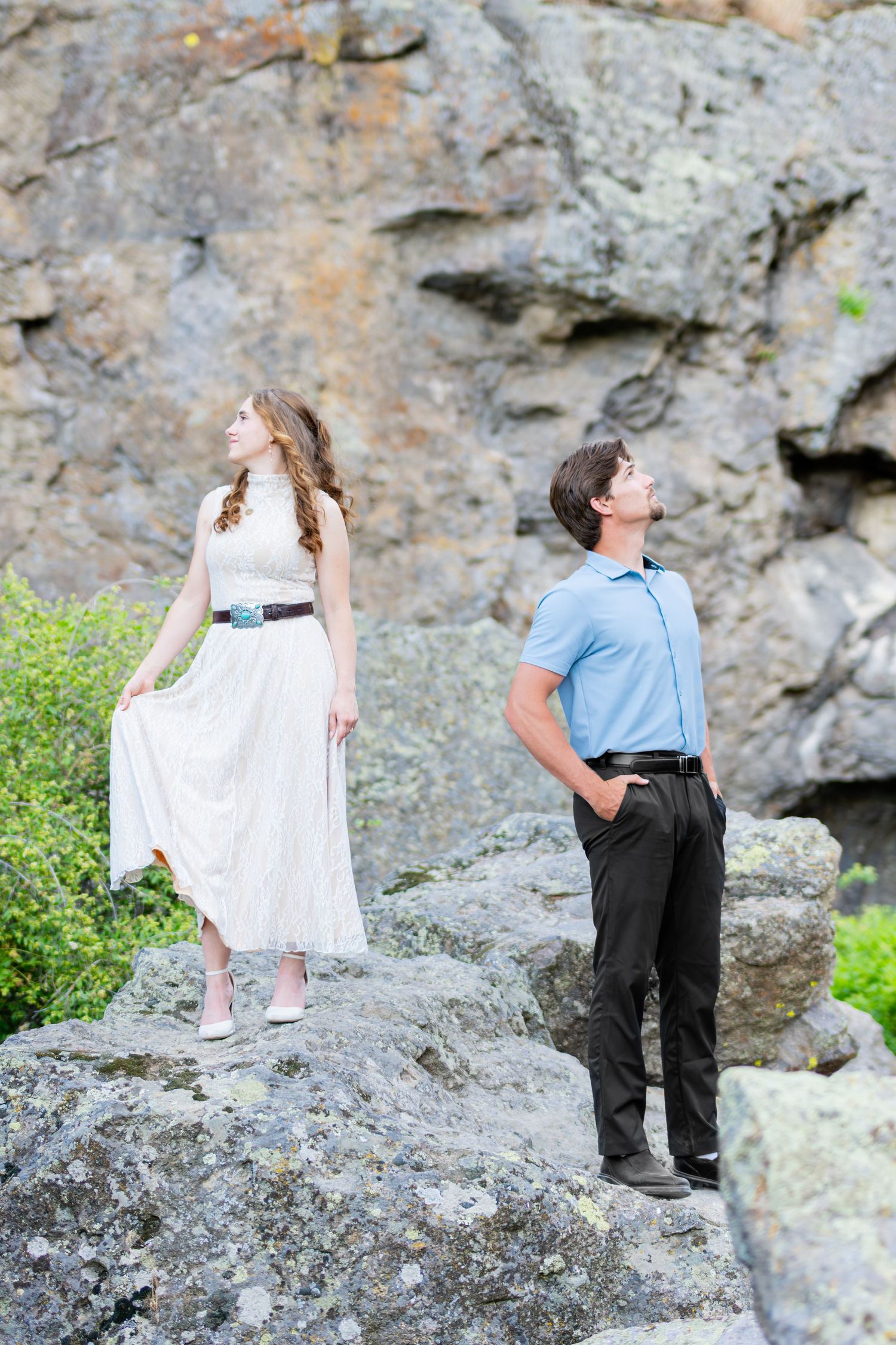 A couple stands on rocky terrain, woman in white dress and man in blue polo gazing upward at a stone cliff backdrop.