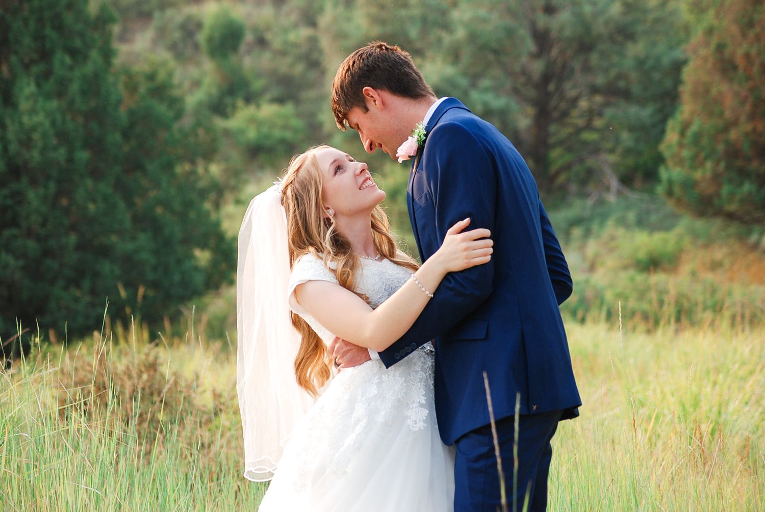 A romantic wedding couple embraces in a sunlit meadow surrounded by lush greenery and natural scenery.