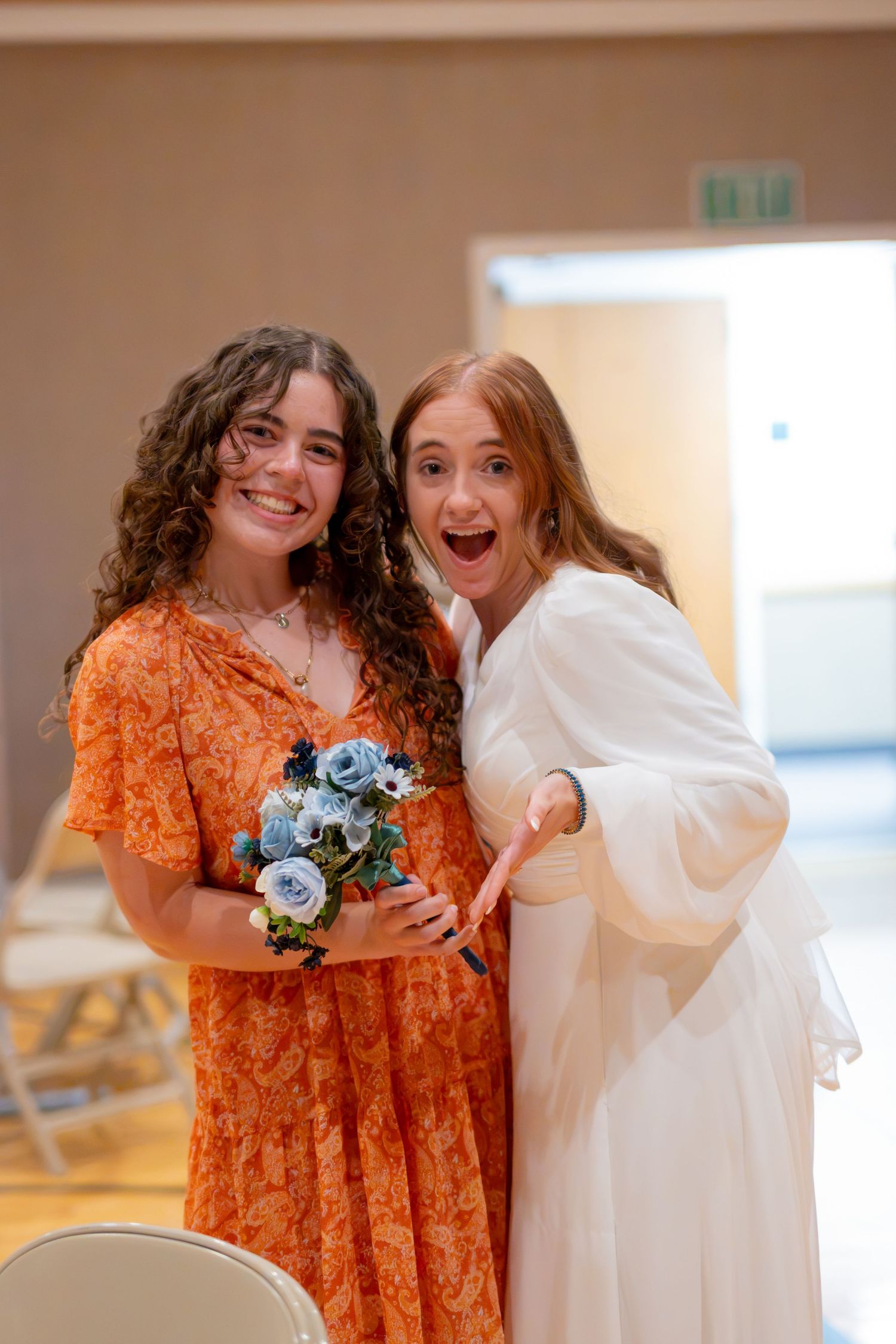 Bride in white dress and bridesmaid in orange floral gown smile holding blue bouquet at wedding reception.