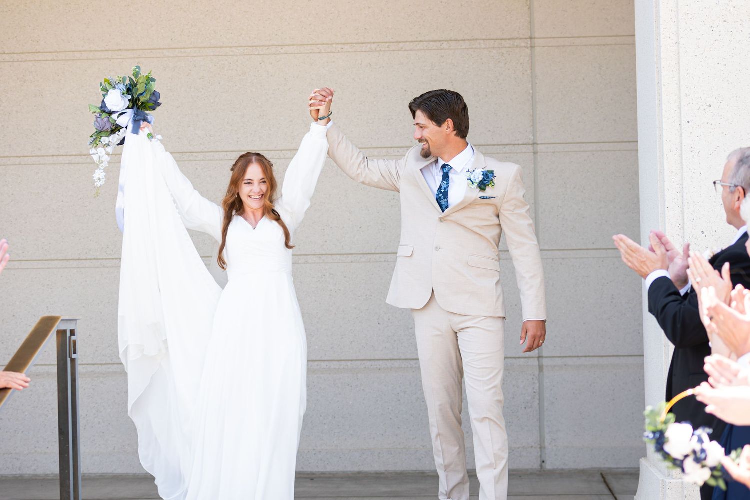 Joyful newlyweds exit wedding venue, bride in white gown holding bouquet aloft, groom in tan suit, guests applauding.