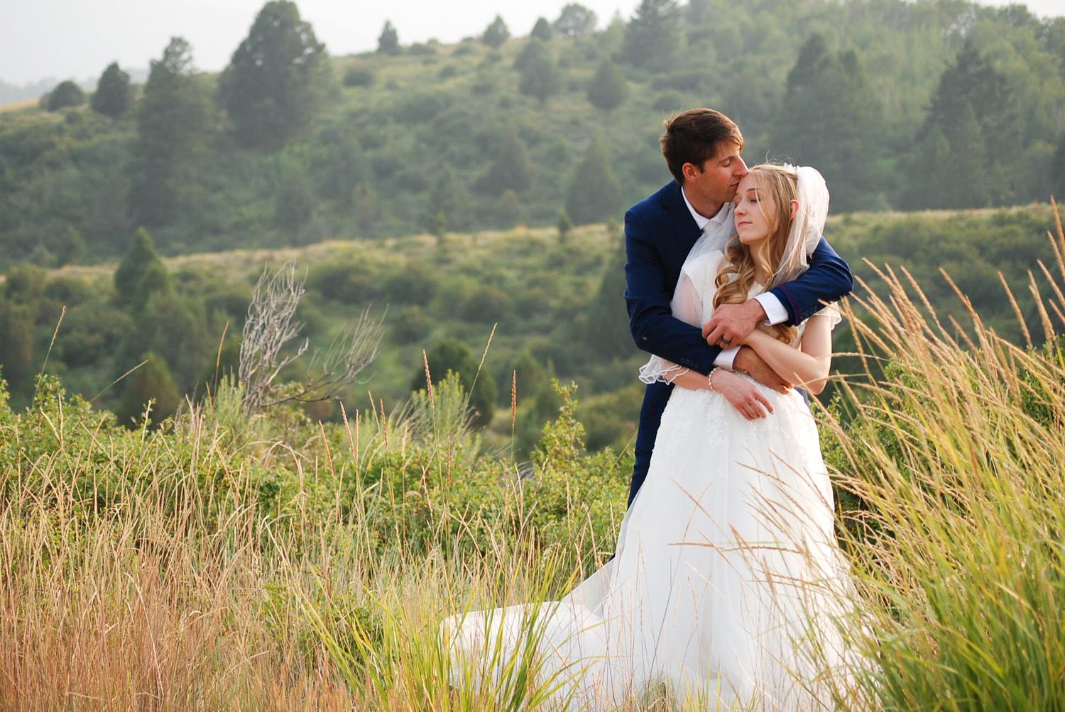 A couple in wedding attire embrace in a scenic field with rolling hills and tall grass during golden hour.