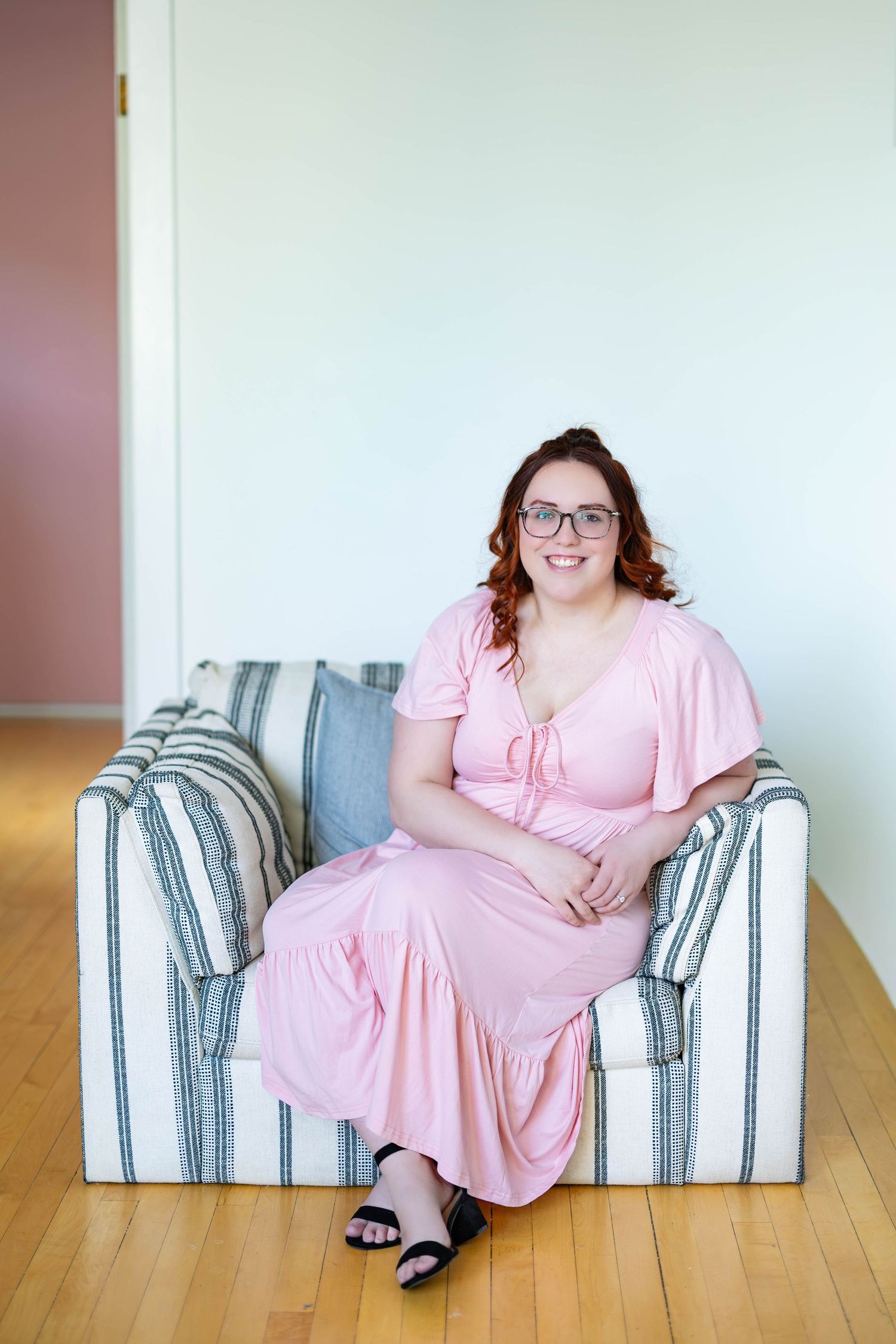 A person in a pink dress poses on a striped armchair in a minimalist room setting.