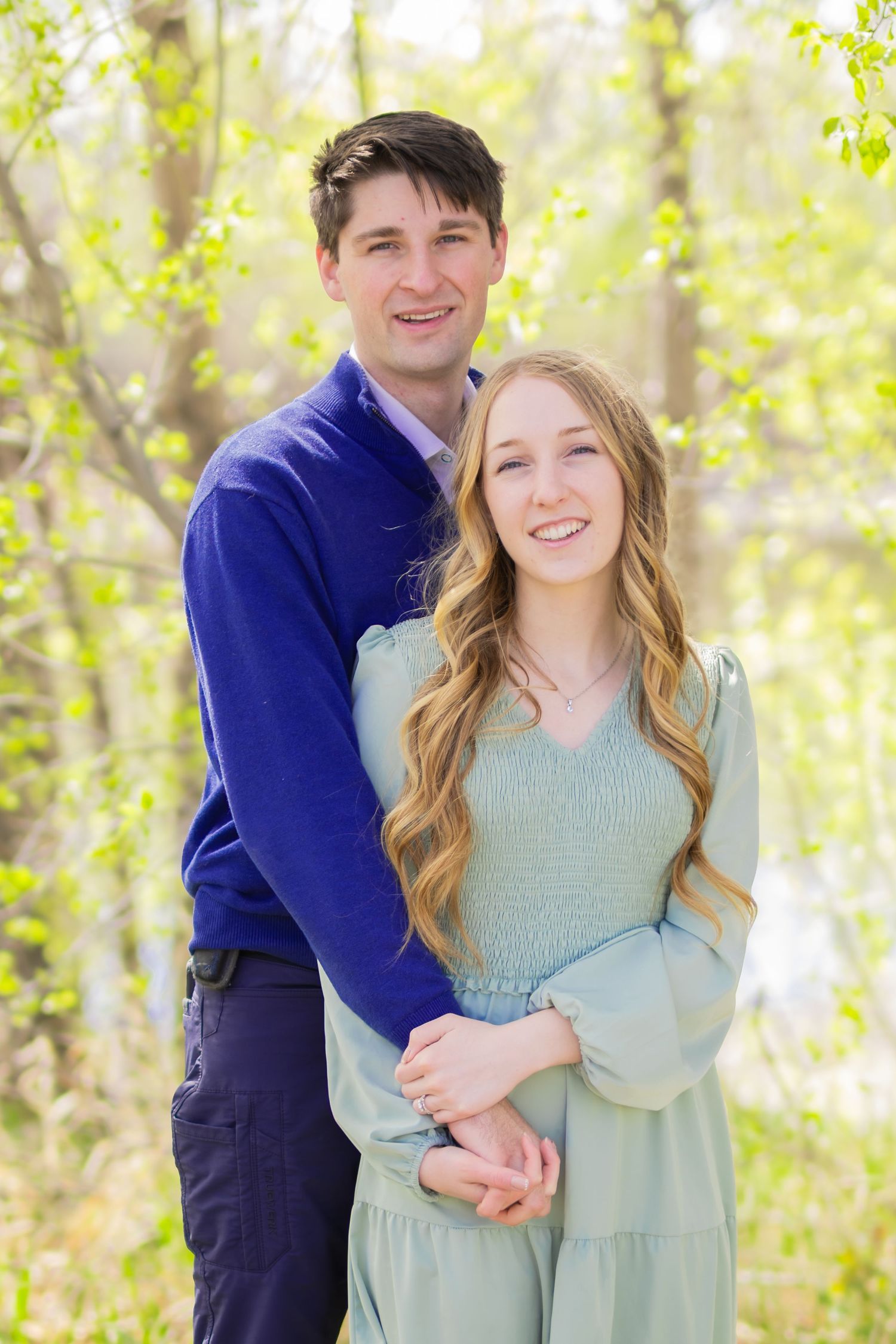 A smiling couple poses outdoors in spring, man in blue sweater embracing woman in mint dress amid green foliage.