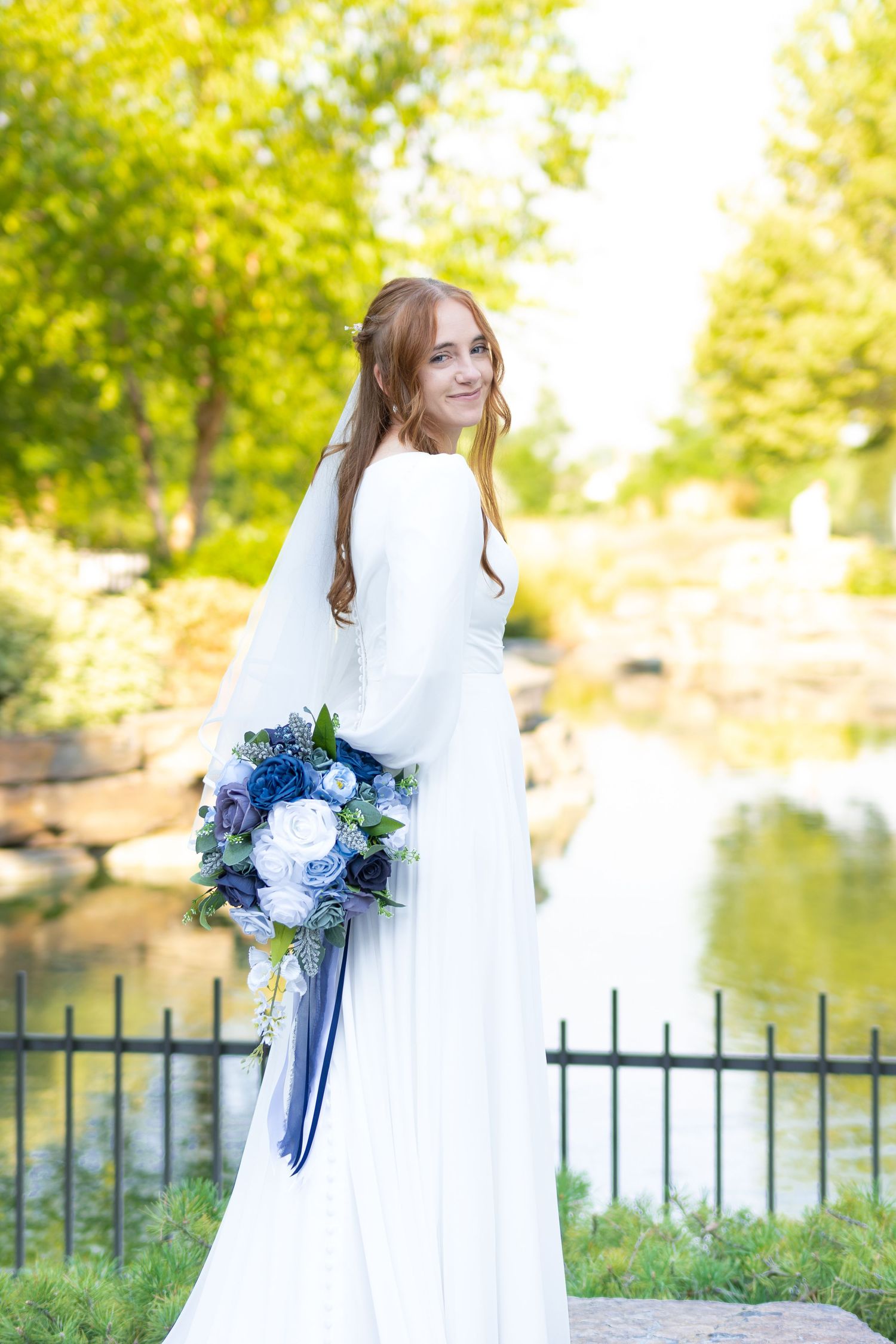 Bride in white long-sleeve gown holding blue and white bouquet, standing near a pond in a green outdoor setting.