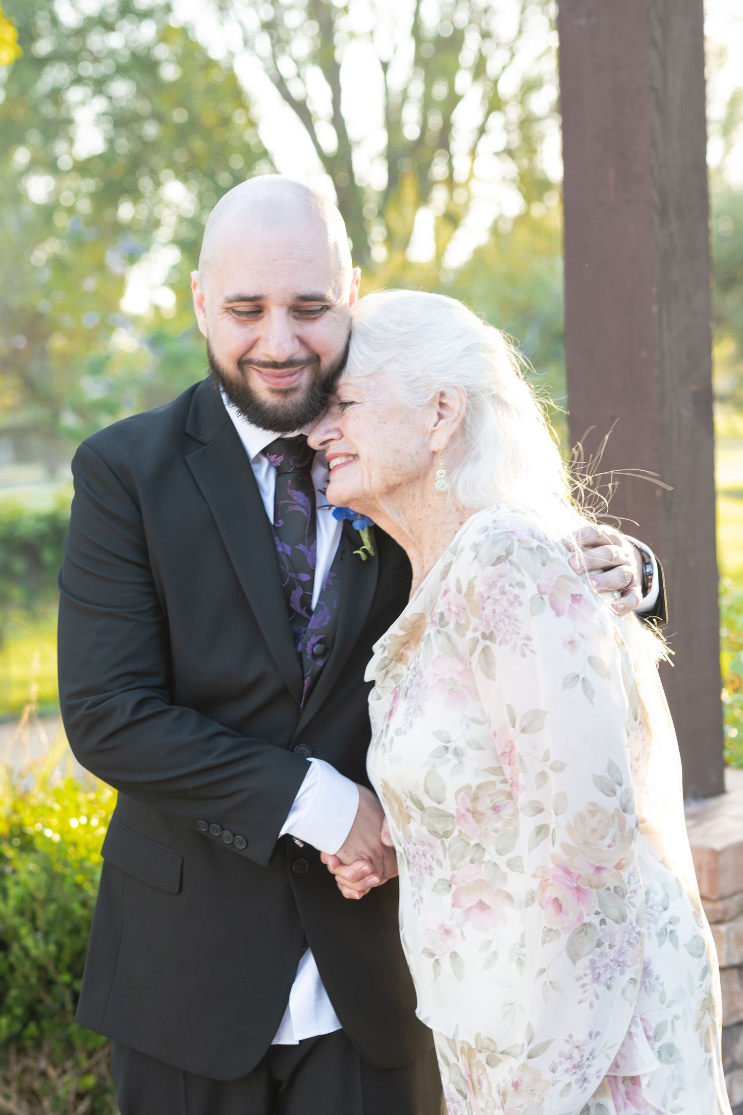 Bald groom in black suit embraces elderly white-haired woman in floral dress outdoors at wedding.
