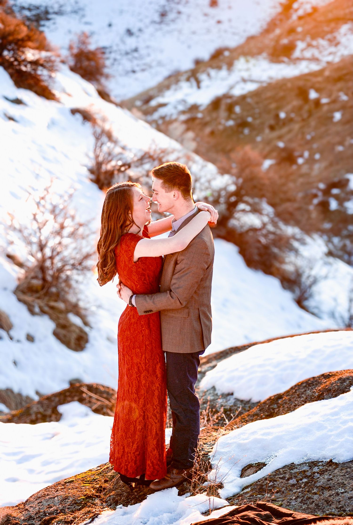 A couple embraces on a snowy mountainside with red rocks and winter landscape in the background.
