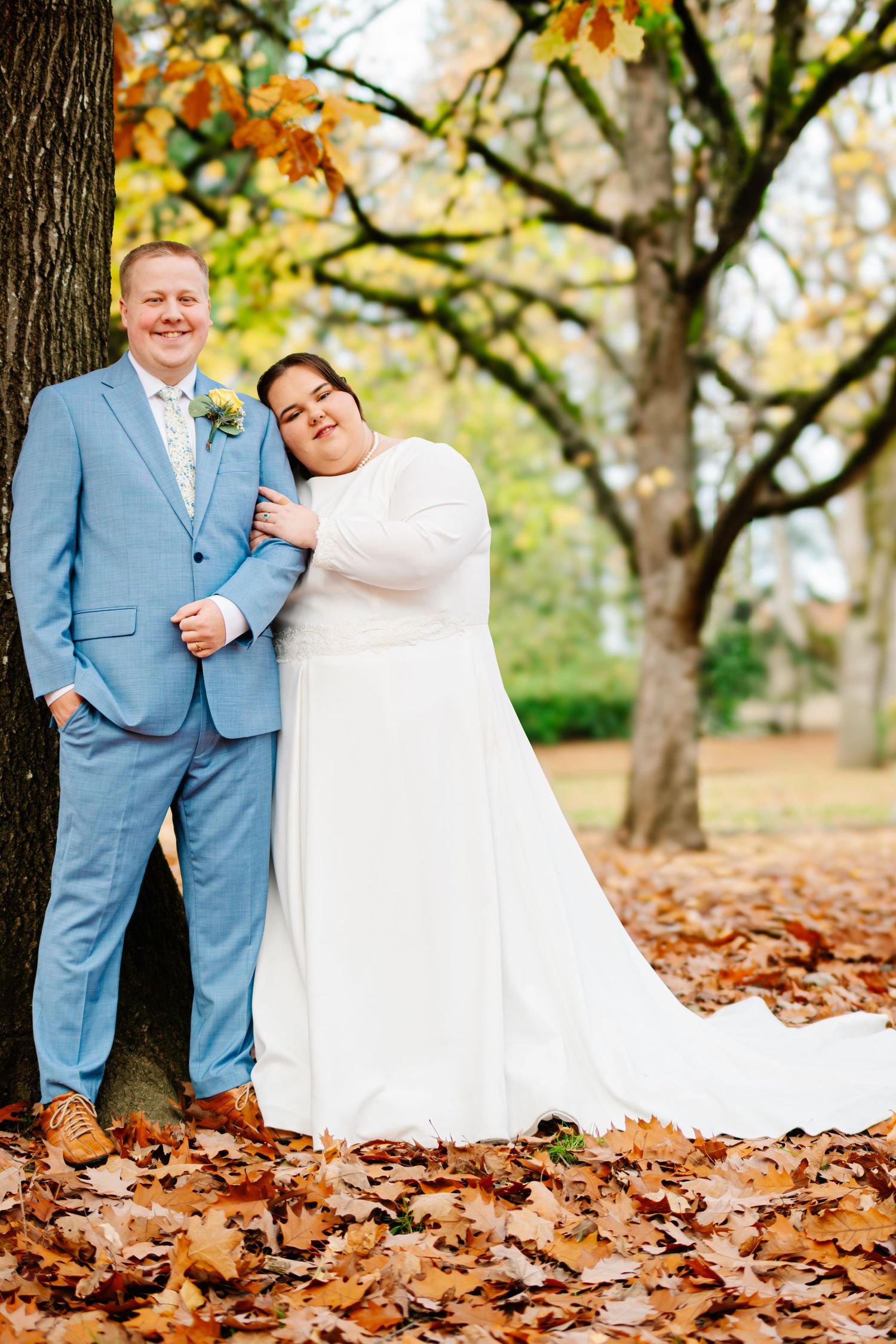 Bride and groom pose among fall leaves by a tree, she in a white gown, he in a light blue suit.