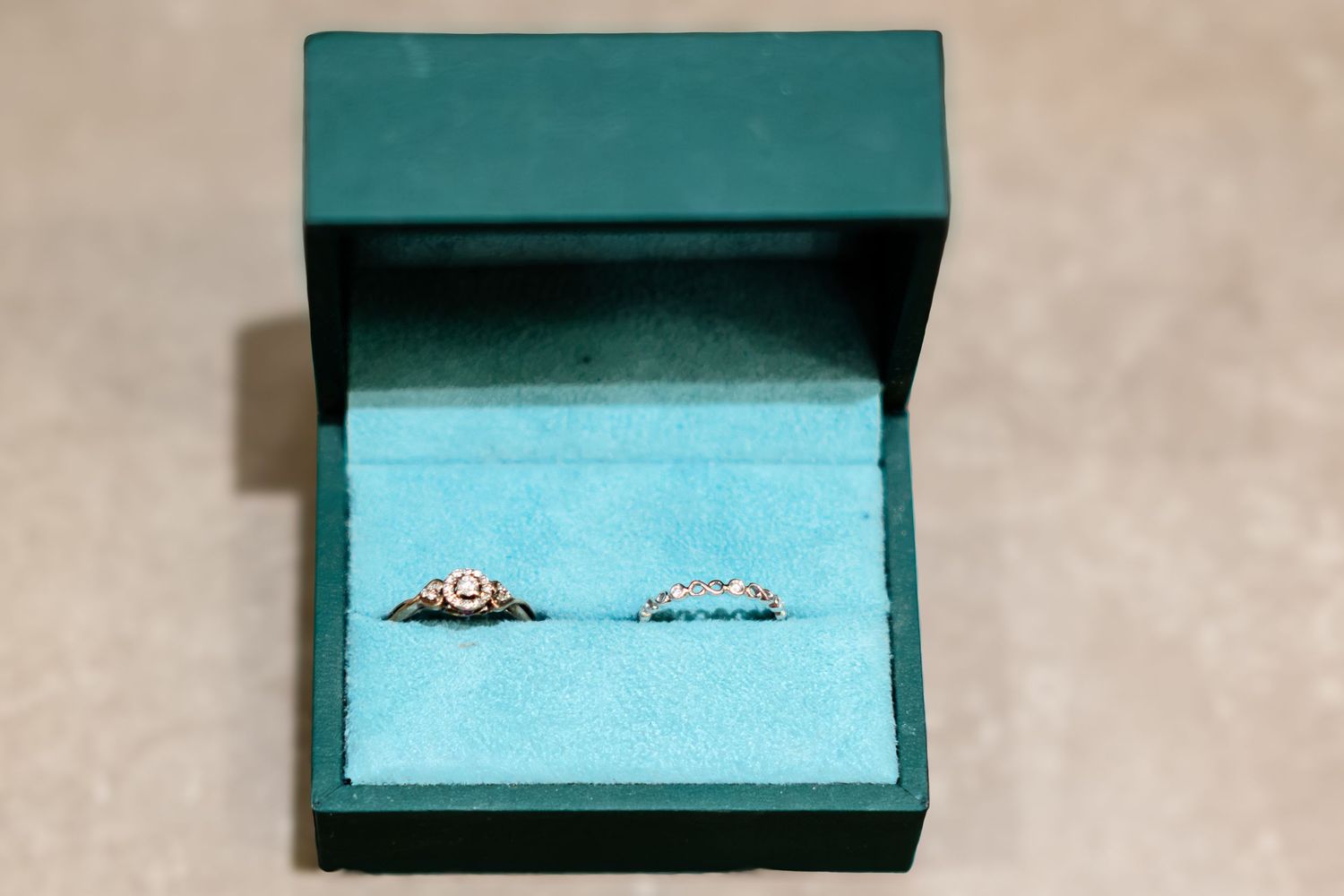 Two delicate diamond rings displayed in an open teal velvet jewelry box with dark green exterior.