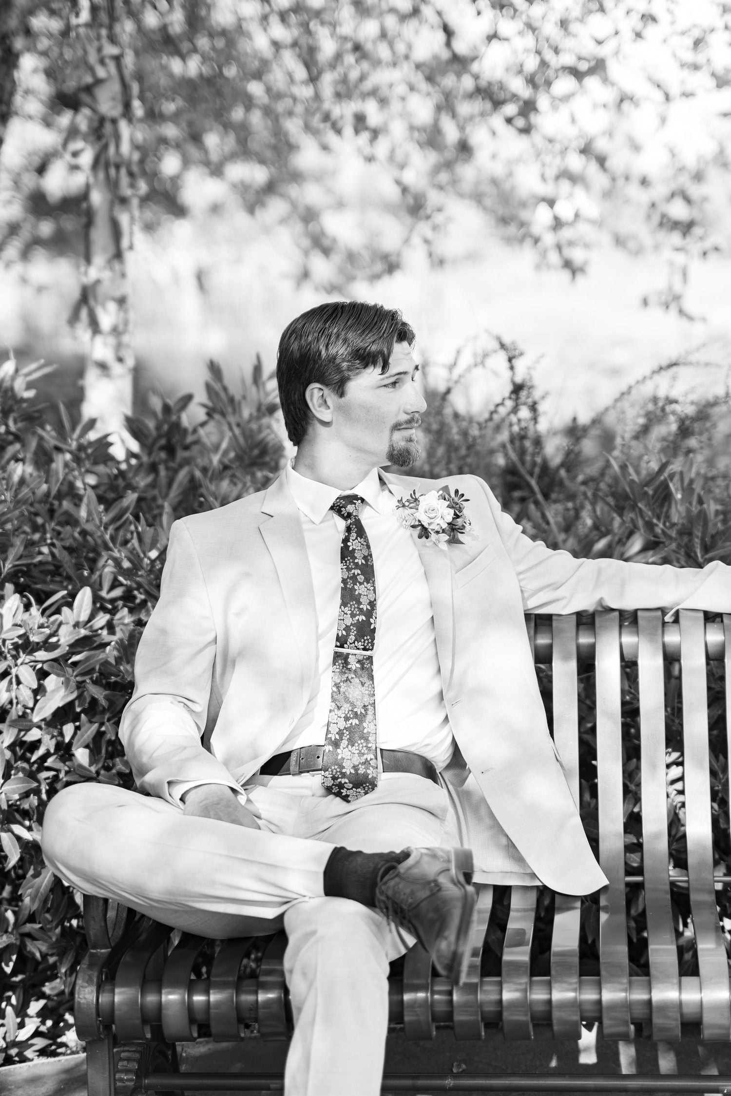 Black and white photo of a groom in a light suit and floral tie sitting on a wooden bench outdoors.