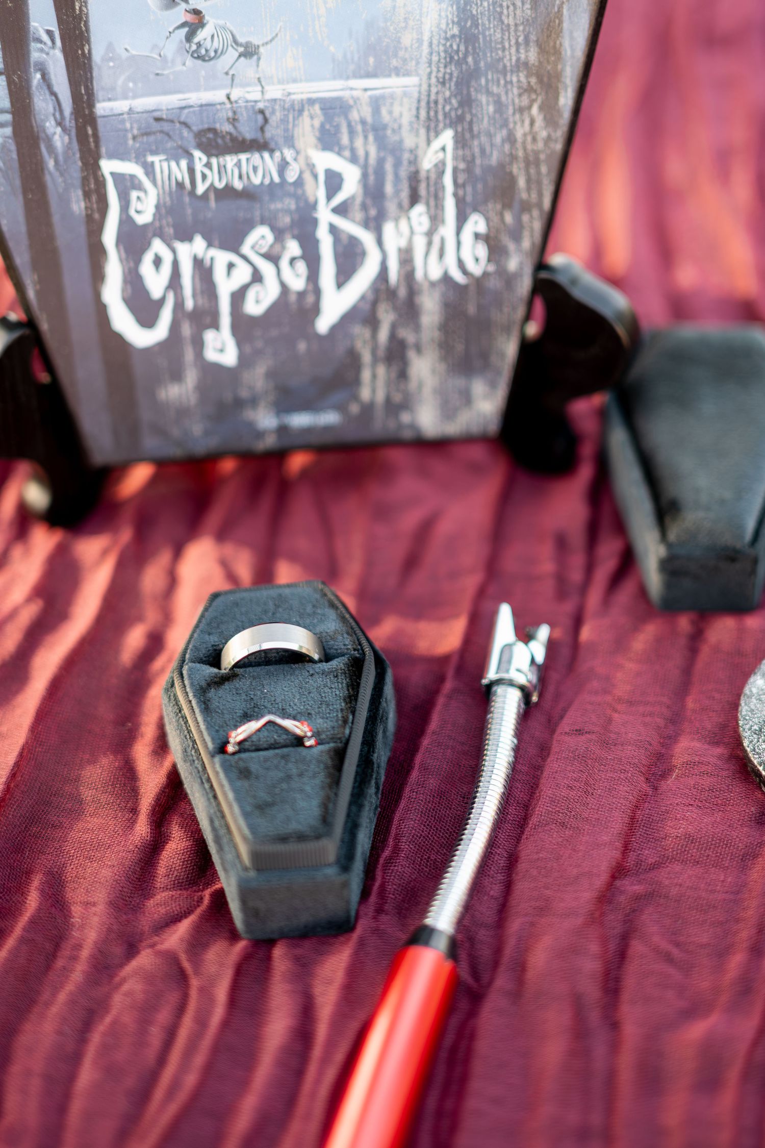A coffin-shaped ring box with wedding bands displayed beside a Tim Burton Corpse Bride box on red fabric.