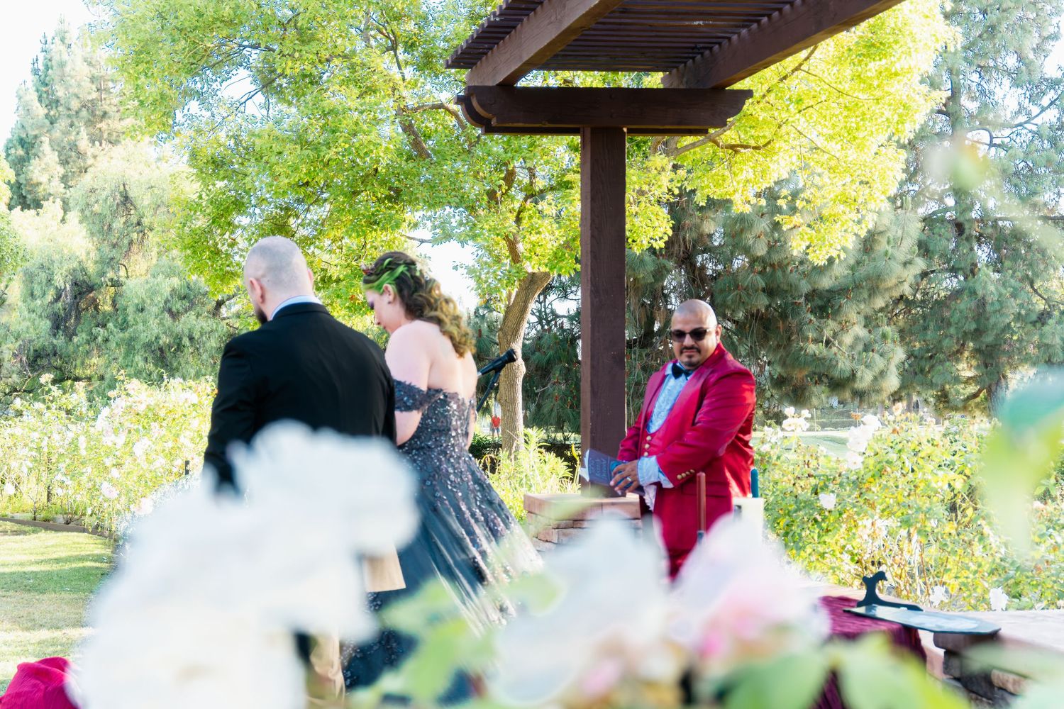 Officiant in red jacket presides over outdoor wedding ceremony under wooden pergola surrounded by lush greenery and flowers.