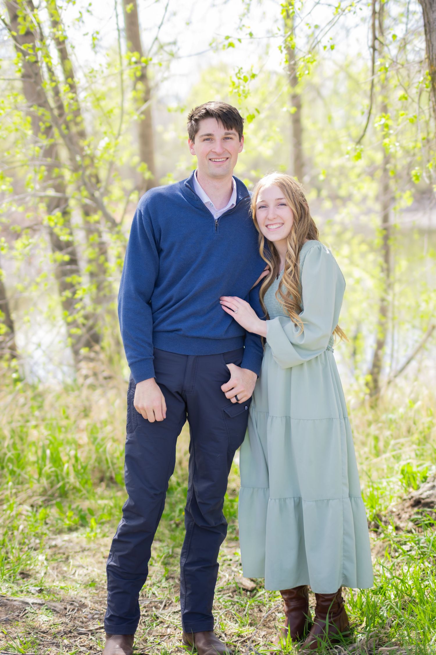 Smiling couple posing outdoors among budding spring trees, he in a navy sweater and she in a sage green tiered dress and brown boots.
