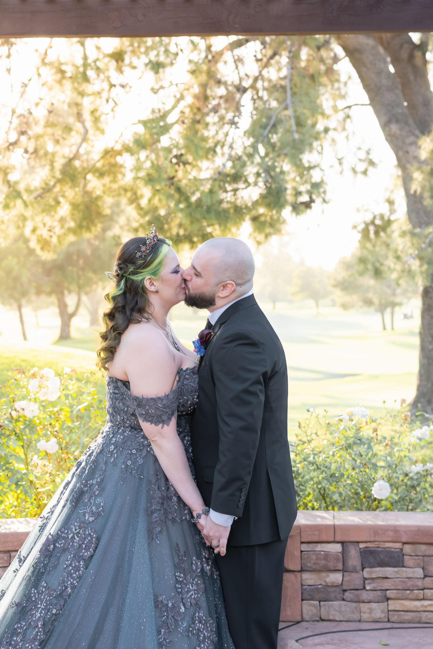 Couple sharing a kiss in wedding attire, bride in gray gown with floral crown, groom in black suit, outdoors.
