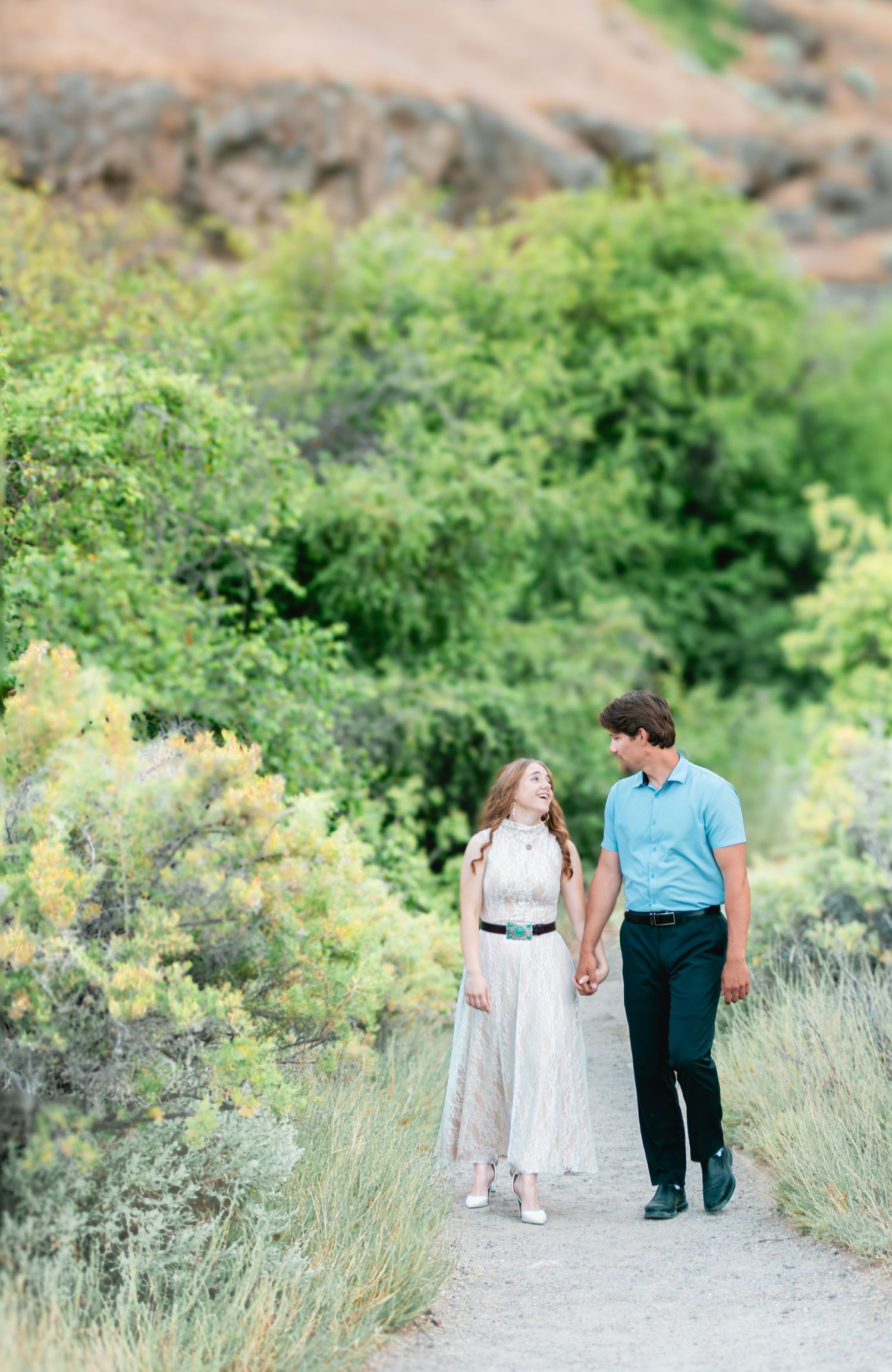 Couple holding hands walking on a scenic path surrounded by lush green bushes and rocky hills.