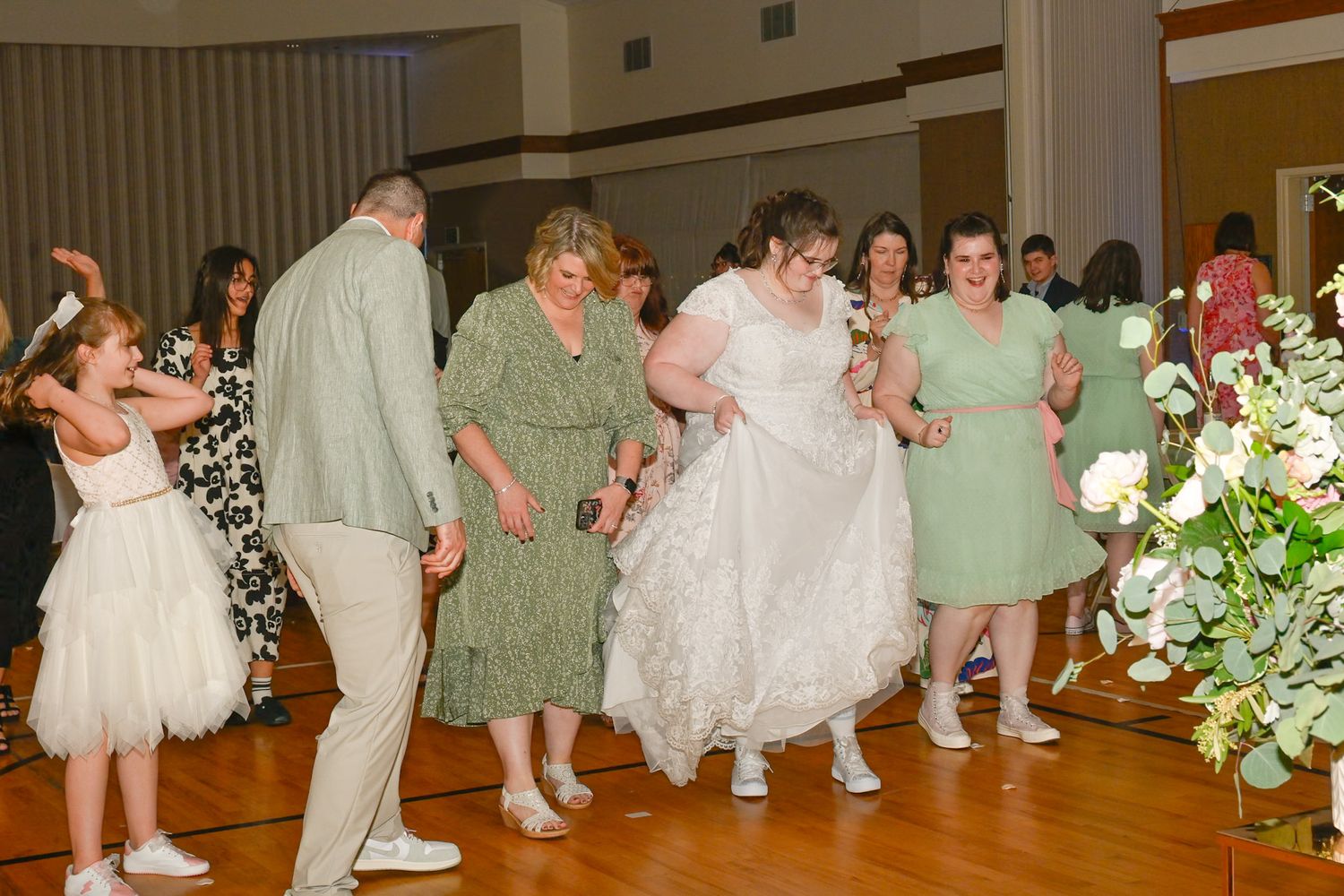 Wedding guests dance together in a line on a wooden dance floor with floral decorations visible in the reception hall.