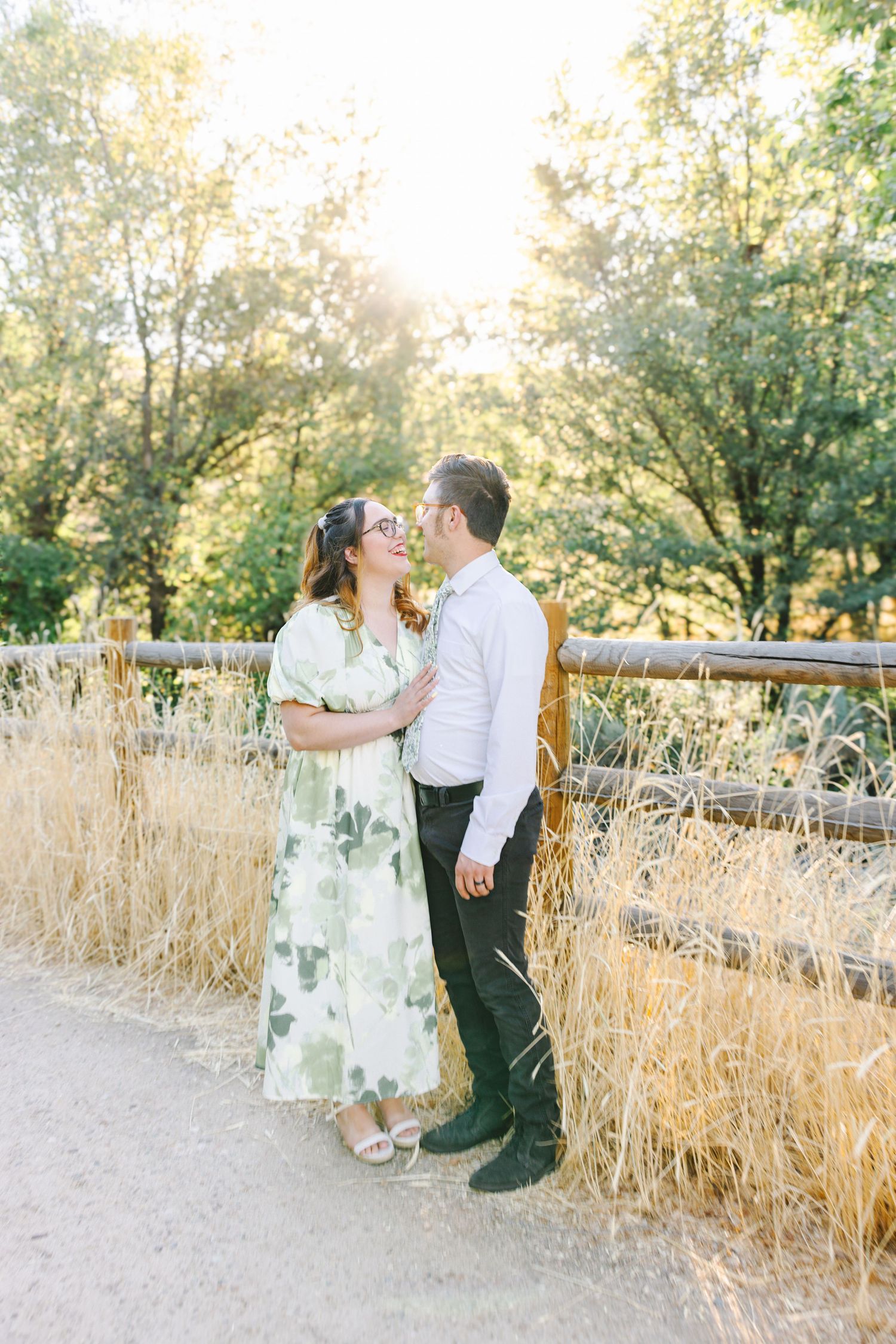 Smiling couple in a floral dress and white shirt embrace beside a wooden fence on a sunlit dirt path surrounded by golden grass.