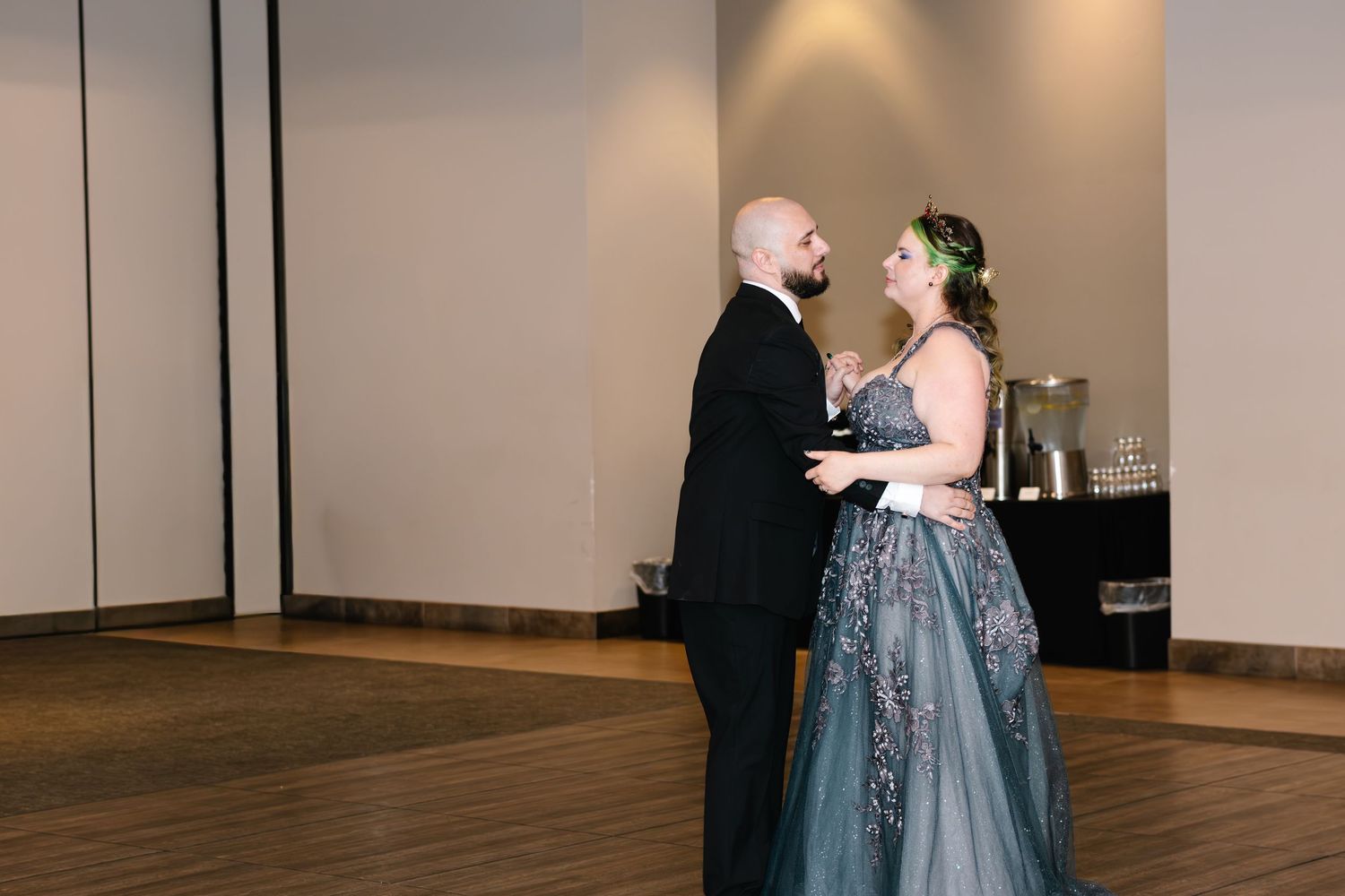 Couple sharing first dance in elegant ballroom, bride in blue-gray gown with floral details, groom in black tuxedo.