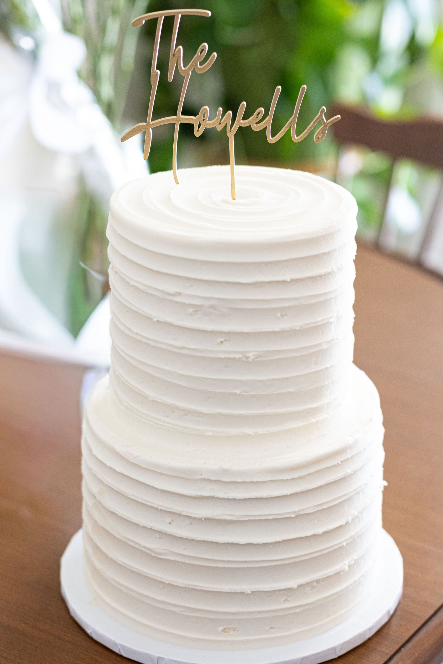 Simple white textured wedding cake with gold script topper on wooden table.