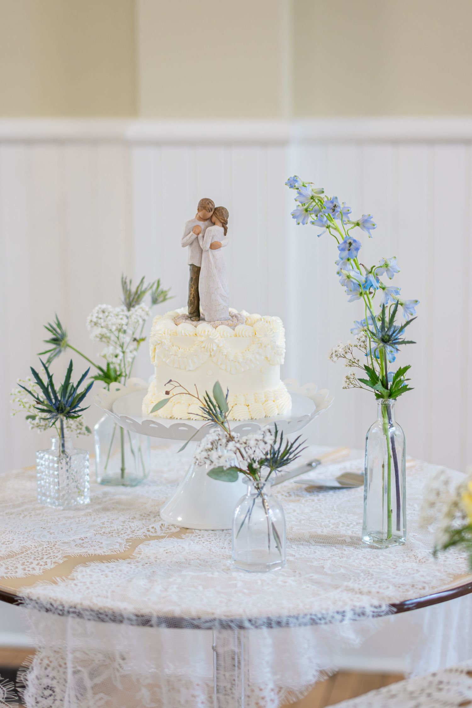 White wedding cake with couple topper on lace table, surrounded by small glass vases with blue and white flowers.