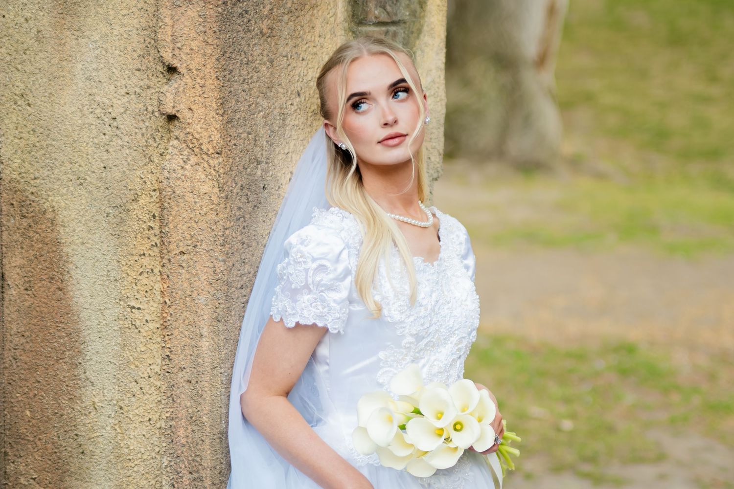 Blonde bride in white lace dress holding white flower bouquet, leaning against stone wall outdoors.