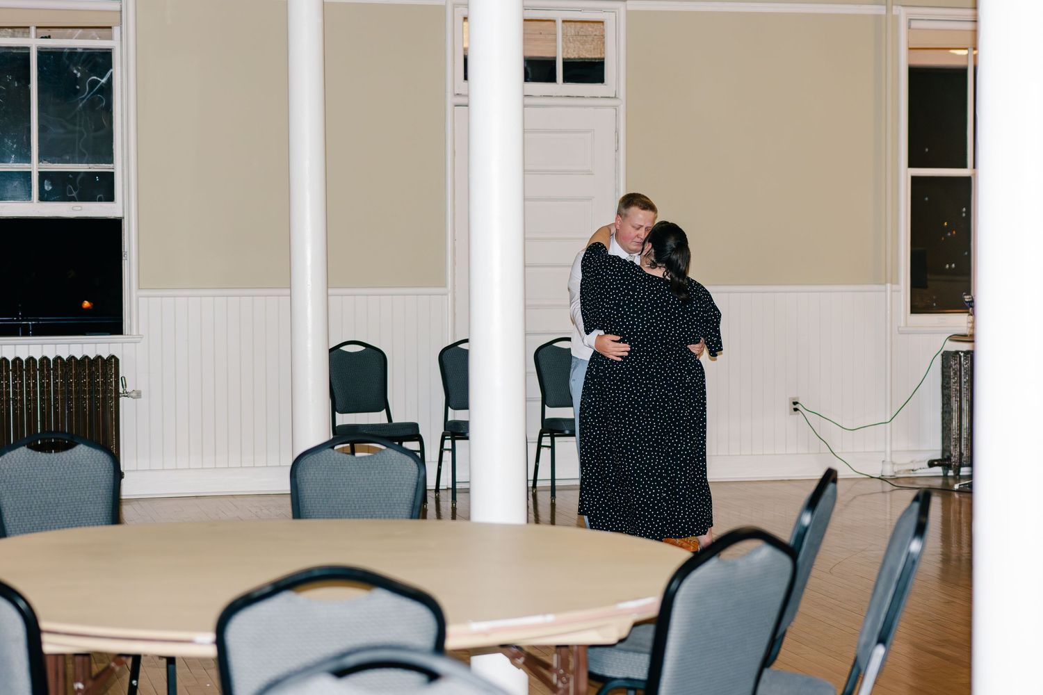 Two people embrace in a meeting room with round tables and blue chairs.
