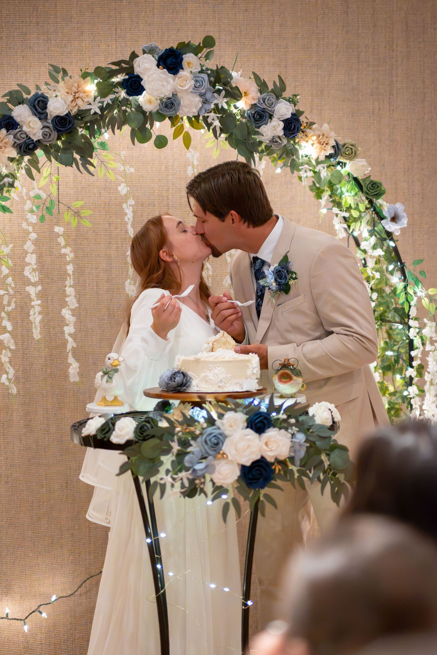 Newlyweds share a kiss while cutting their wedding cake under a floral arch with blue and white flowers.